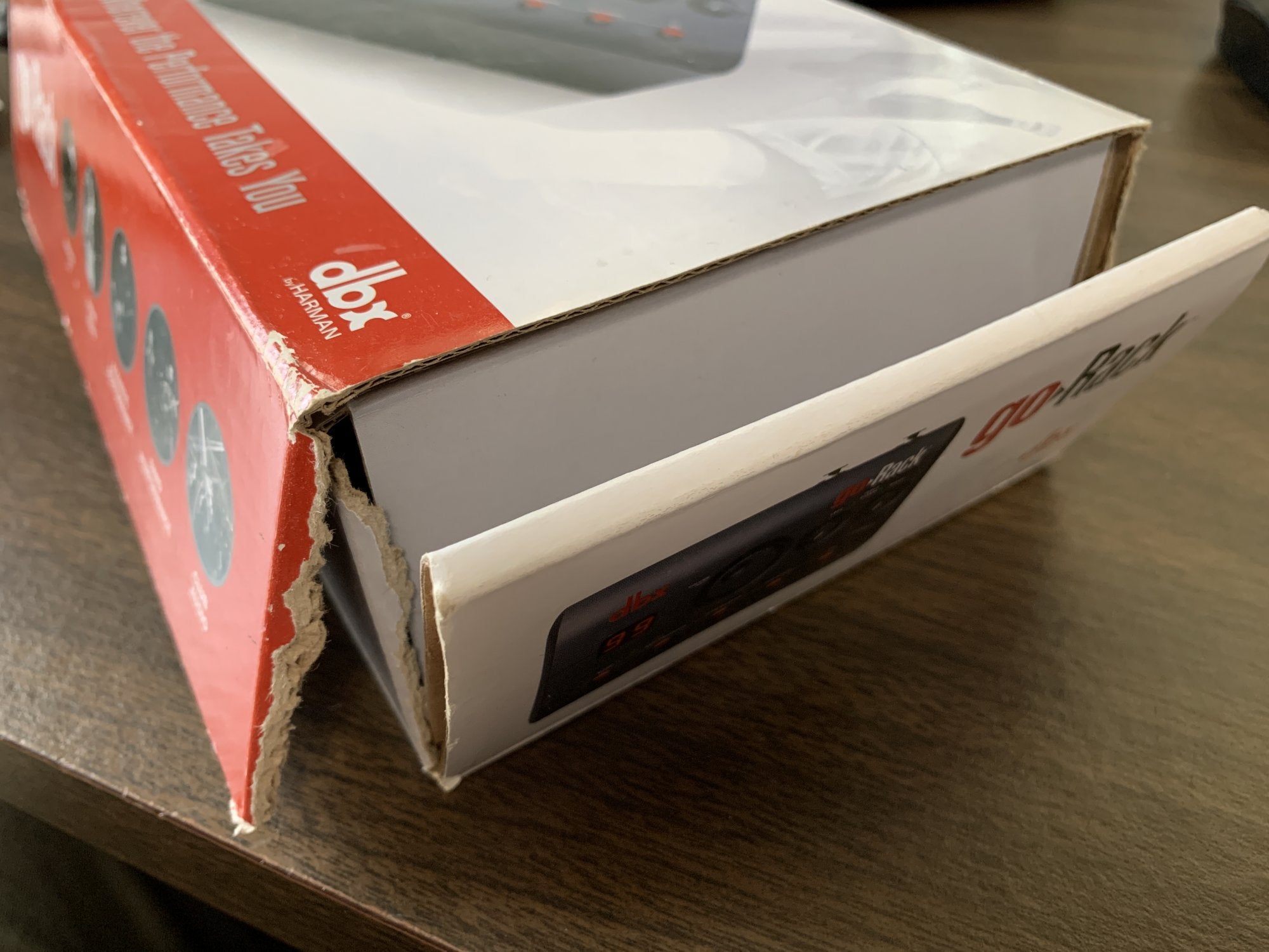A close-up of a partially opened box with torn edges reveals the white and red packaging for the (Mint) DBX goRack Performance Processor by DBX, resting on a brown wooden surface.
