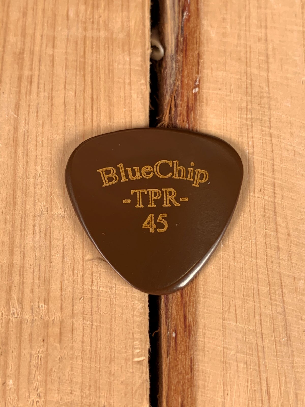 A precision-machined BlueChip Picks (TPR45) guitar pick from BlueChip rests on wood, featuring "BlueChip TPR 45" engraved in gold and a narrow vertical gap beneath it.