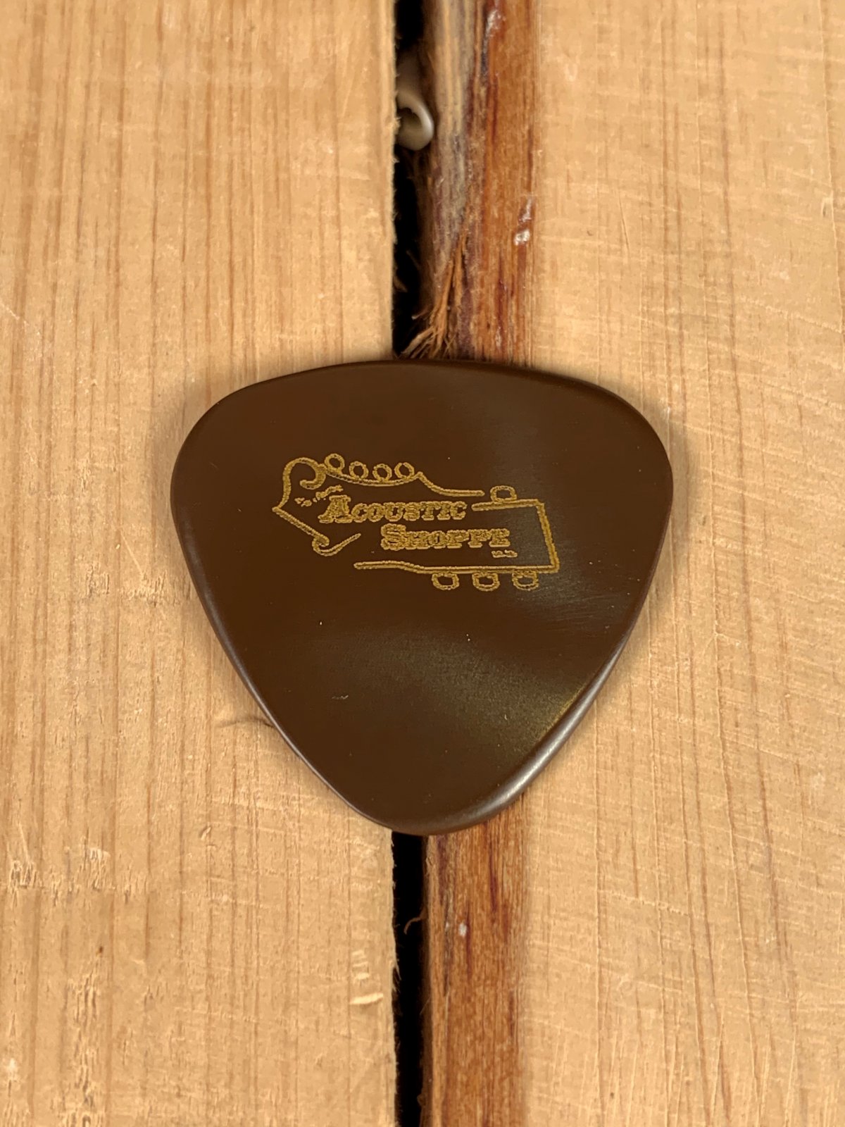 A BlueChip Picks (TPR45) by BlueChip, a brown precision-machined guitar pick featuring "Acoustic Shore" and a gold guitar headstock design, rests on a light wooden surface with visible board gaps.