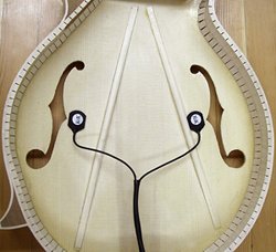 The image shows the inside of a mandolin with a K&K Mandolin Twin Internal Pickup by K&K Sound attached near the f-shaped sound holes, along with visible interior bracing and wooden ribs.