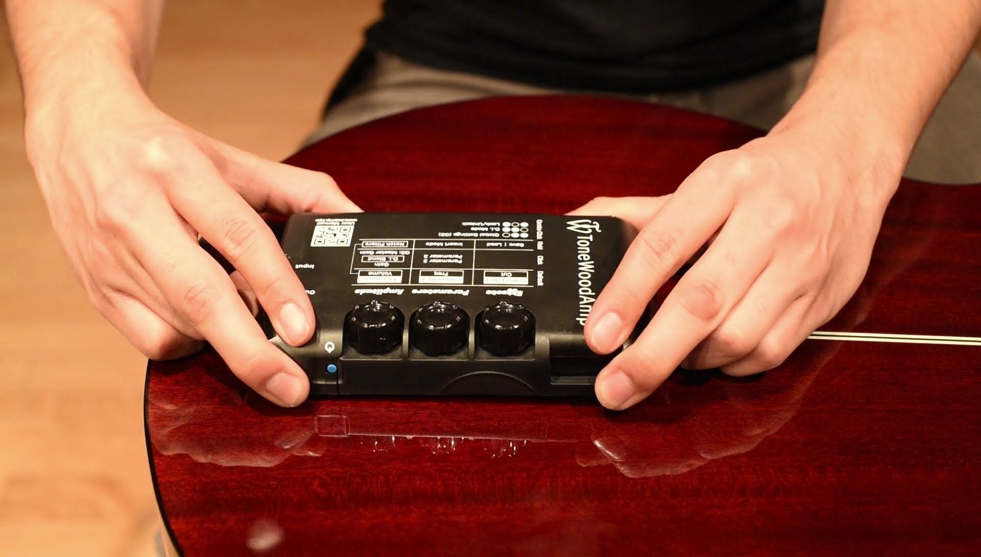 Close-up of hands installing a HONZ Technologies ToneWoodAmp Solo Left Handed multi-effect processor with control knobs onto the glossy, reddish-brown body of an acoustic guitar.