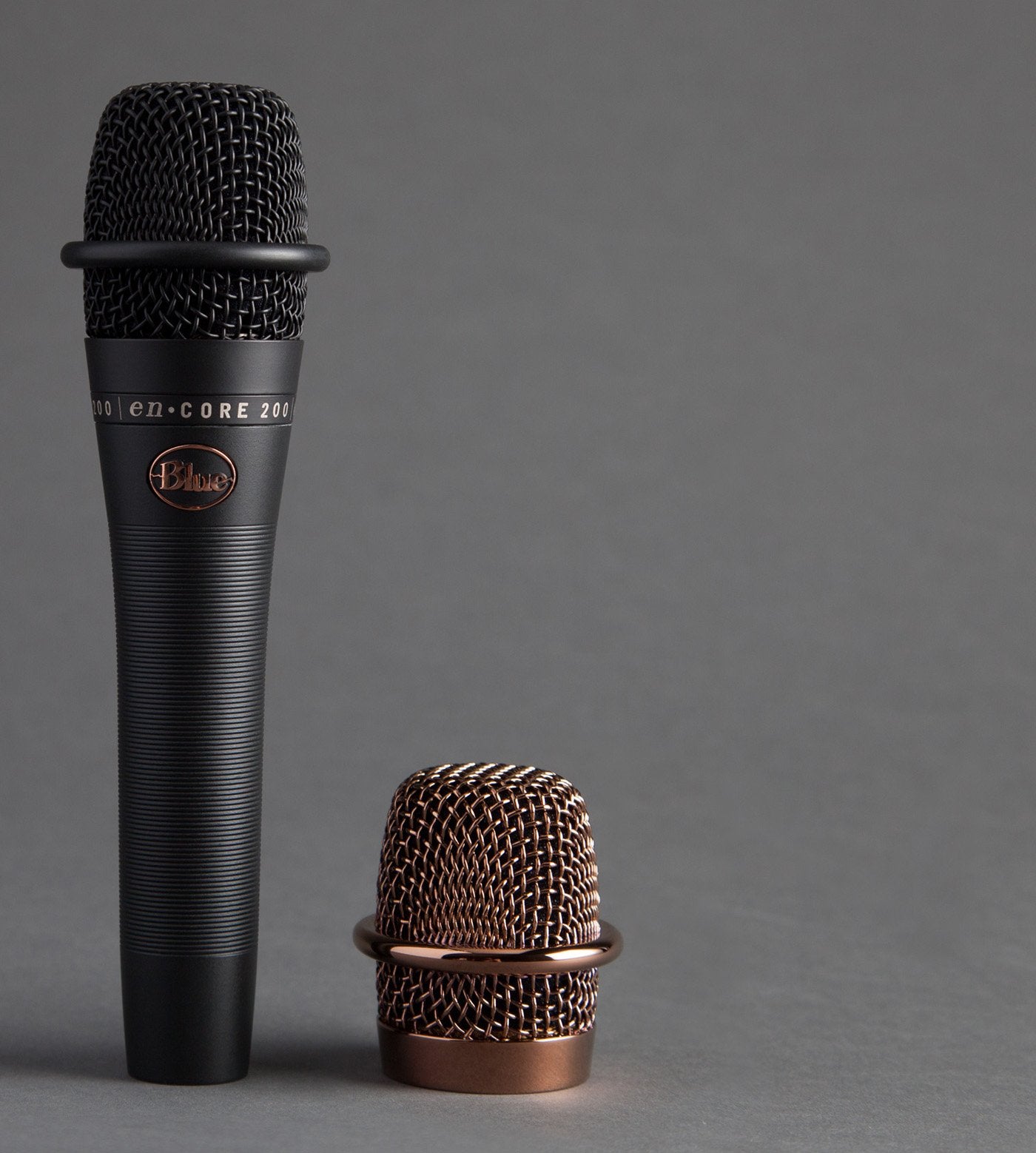 A Blue Microphones Blue Encore 200 Dynamic Microphone (HL00211520) in black stands upright beside its separate bronze mesh grille on a gray surface, with a plain gray background.