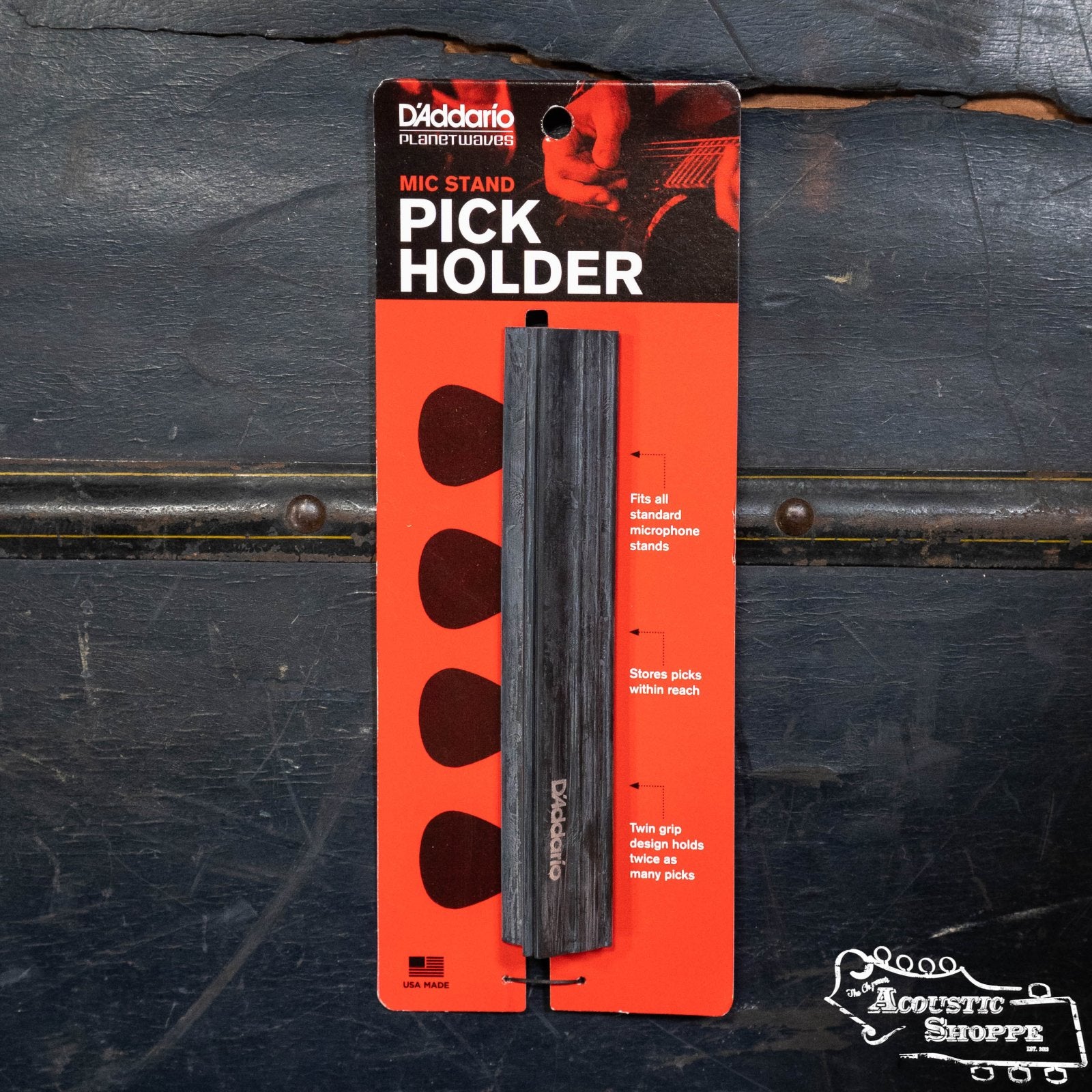 A D'Addario Mic Stand Pick Holder PW-MPH-01, in orange and black packaging, is displayed on a dark wooden surface. The accessory has four circular pick storage areas and features the D'Addario brand.