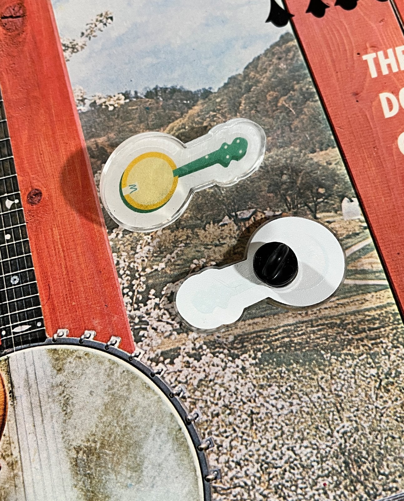 Two puzzle pieces rest on a jigsaw featuring the Mark A Montgomery Yellow Banjo Acrylic Lapel Pin—one piece shows a yellow banjo illustration, the other is white with a black knob—celebrating acoustic instruments.