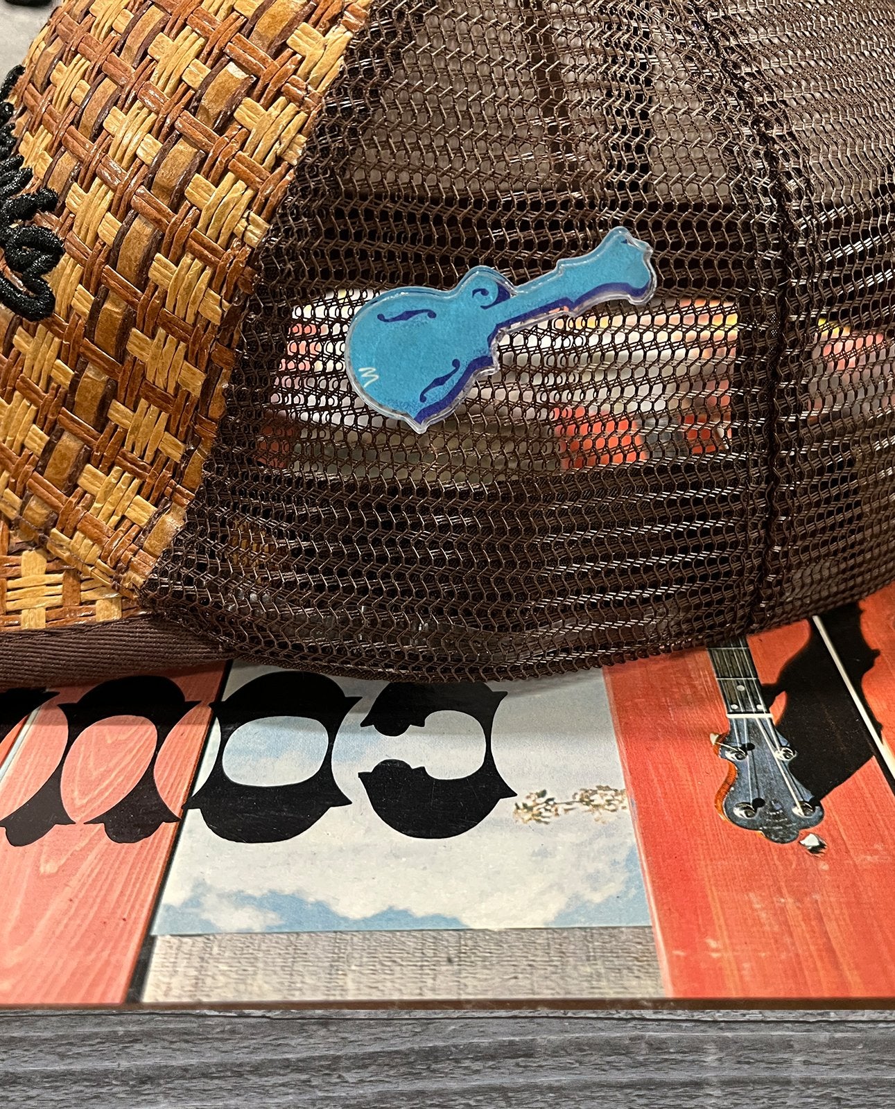 A brown mesh trucker hat with a Blue Mandolin Acrylic Lapel Pin by Mark A Montgomery rests on a vibrant surface featuring guitar illustrations—an ideal accessory for fans of boutique brands and acoustic instruments.