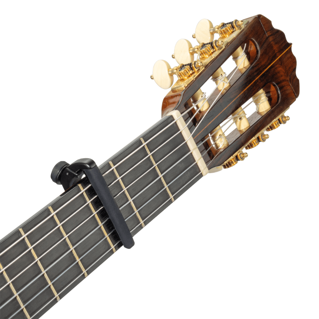 Close-up of a classical guitar's headstock and upper neck with golden tuning pegs. A D'Addario Pro Plus Guitar Capo - Black, featuring FlexFit technology, is clamped across the fretboard, pressing down the strings.