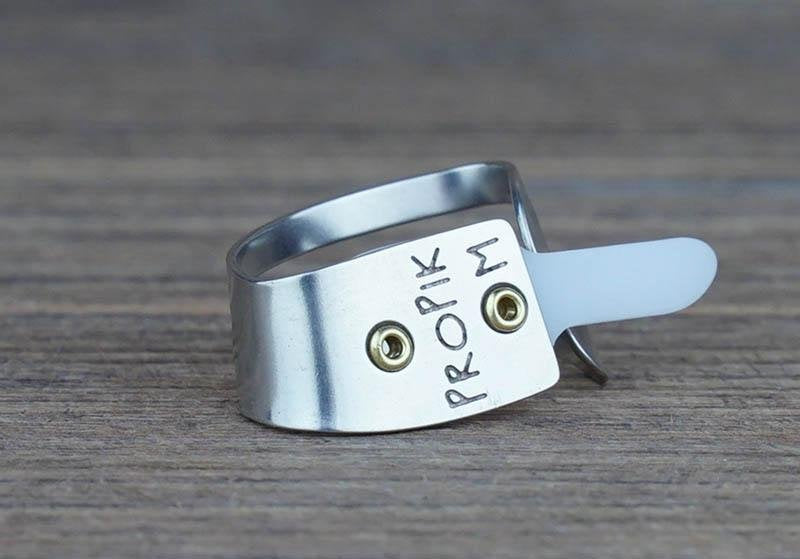 A Deering Banjos ProPik Quik-Pik Thumb Pick Medium (11-206-3013), featuring an adjustable metal band with a plastic tip secured by two small screws, rests on a wooden surface.