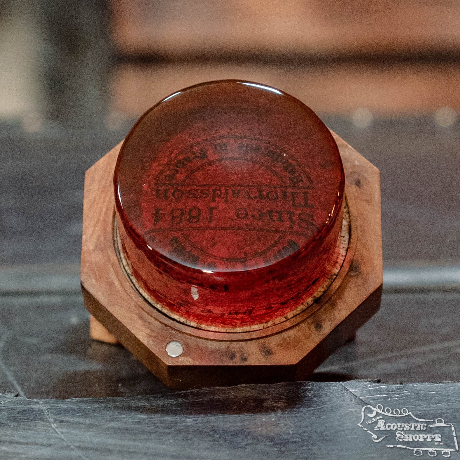 A close-up of Sartory Rosin, a red, round rosin by Sartory, sits on a wooden hexagonal base. Since 1881 and other text appear inside the glass. The Acoustic Shoppe logo is visible in the bottom right corner.