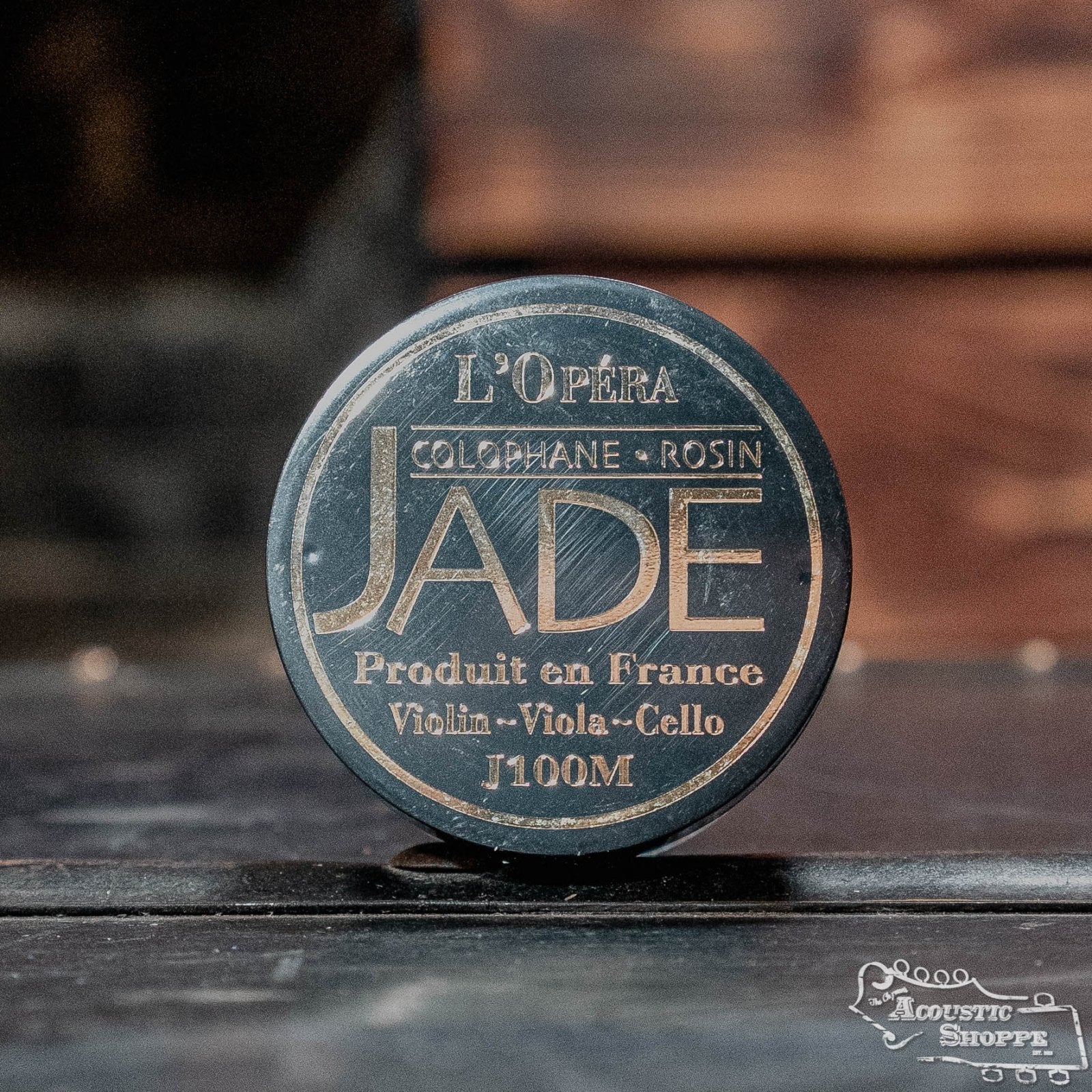 A round tin of Jade Violin/Viola/Cello Rosin by Jade rests on a wooden surface with a blurred background—ideal as premium rosin for violin, viola, or cello.
