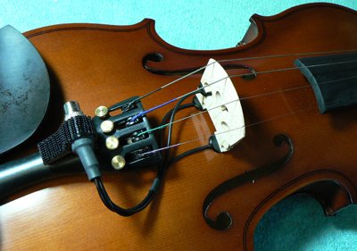 Close-up of a violin with a K&K Violinissimo Pickup by K&K Sound attached near the bridge, showing strings, chinrest, and fine tuners on its wooden body. A textured teal background highlights the pickup’s balanced violin sound.