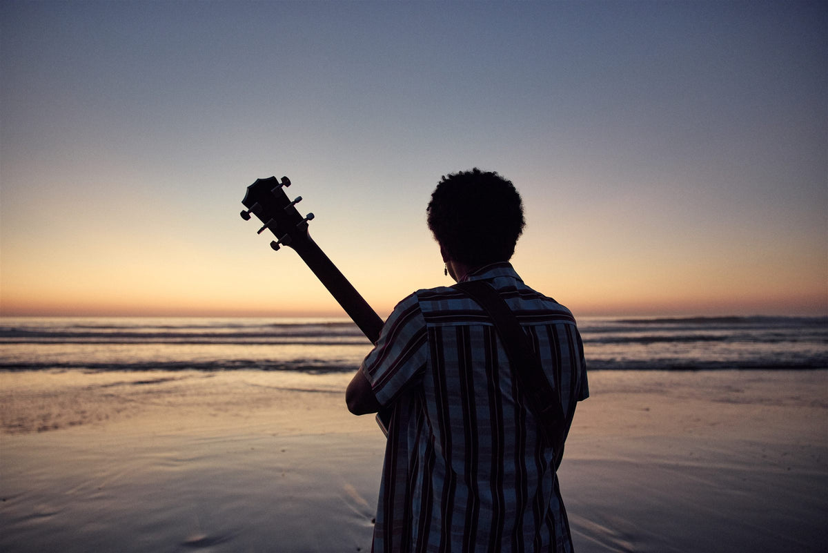 Taylor 300 Series lifestyle photo — musician on a beach at sunset with a Taylor 314ce, The Acoustic Shoppe