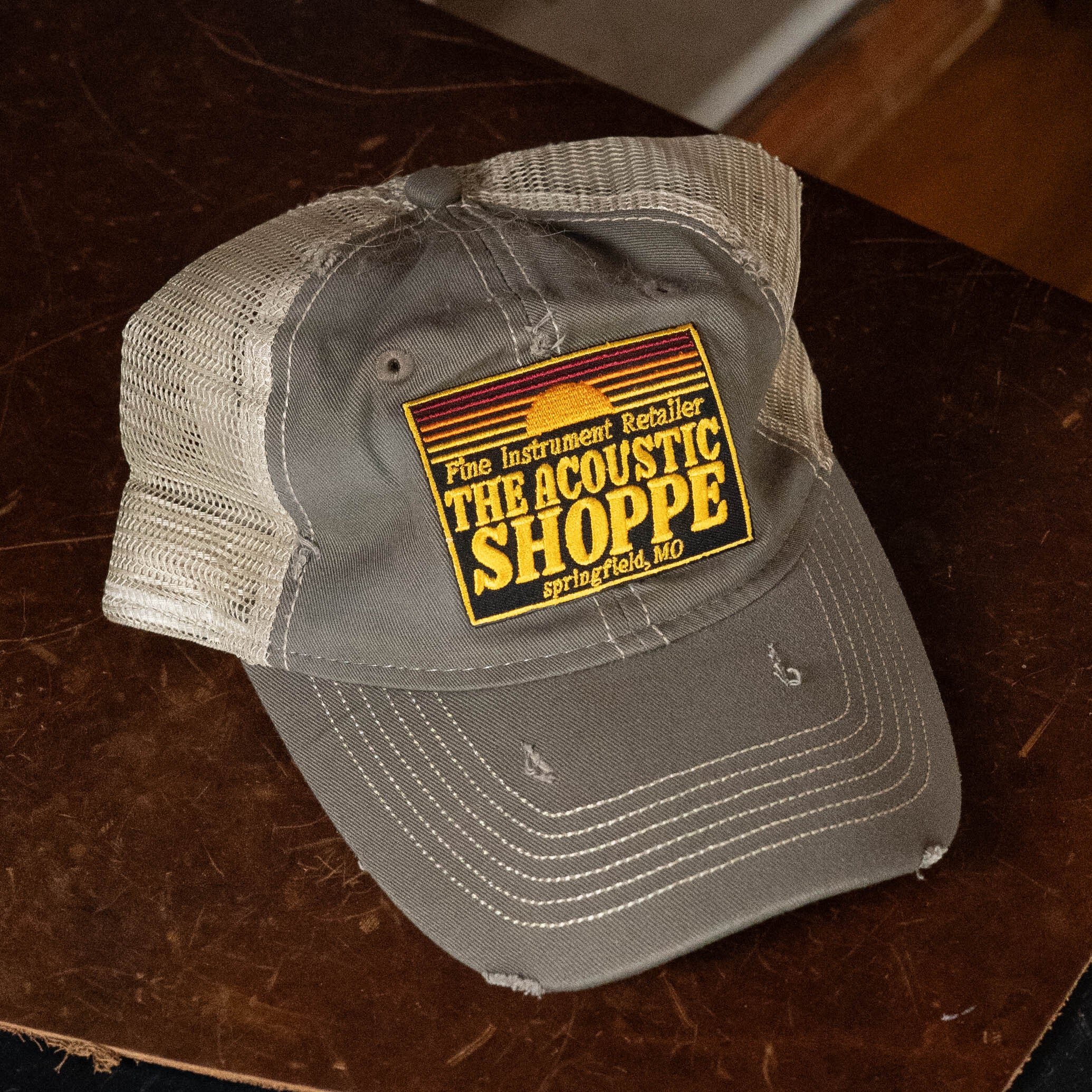 The Acoustic Shoppe Patch Cap Grey by Amplify Screen Printing & Promotions, featuring a sunset patch and gray and beige mesh design, rests on a brown wooden surface.
