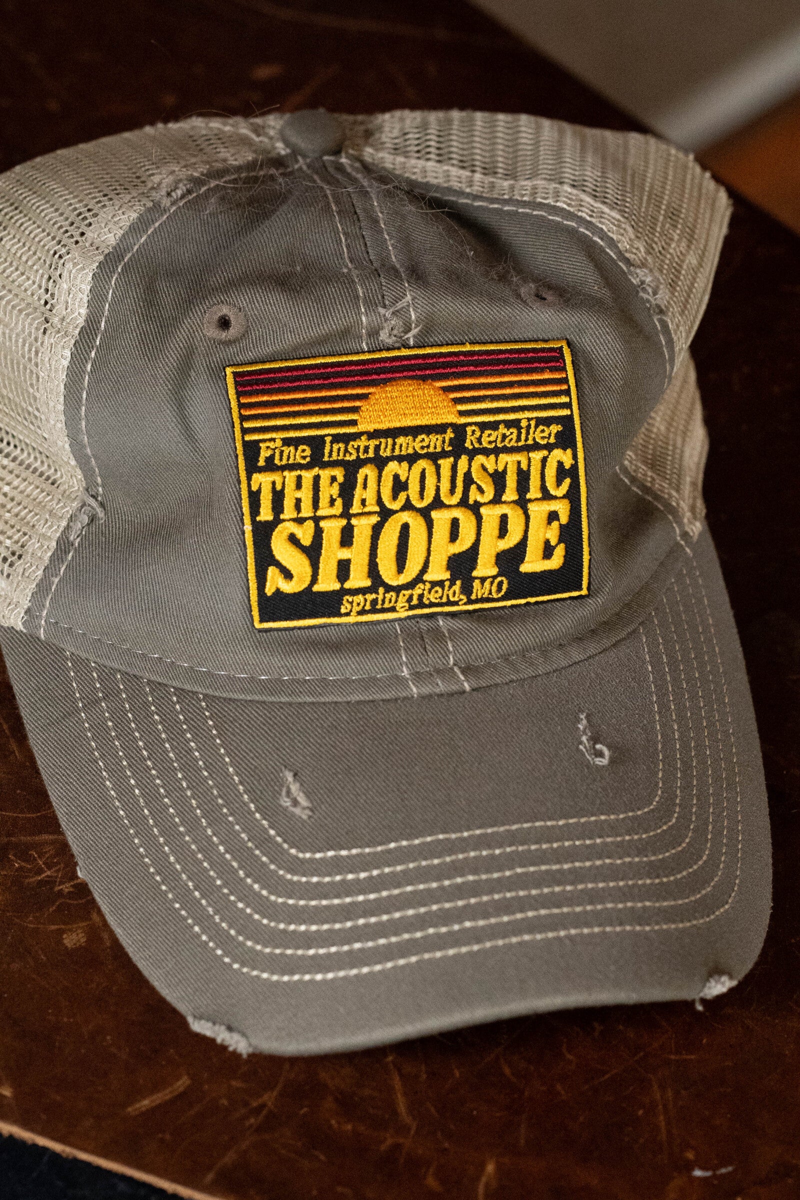 The Acoustic Shoppe Patch Cap Grey by Amplify Screen Printing & Promotions—featuring gray/white mesh, yellow stitching, and a patch with "Fine Instrument Retailer THE ACOUSTIC SHOPPE, Springfield, MO"—rests on a dark wooden surface.