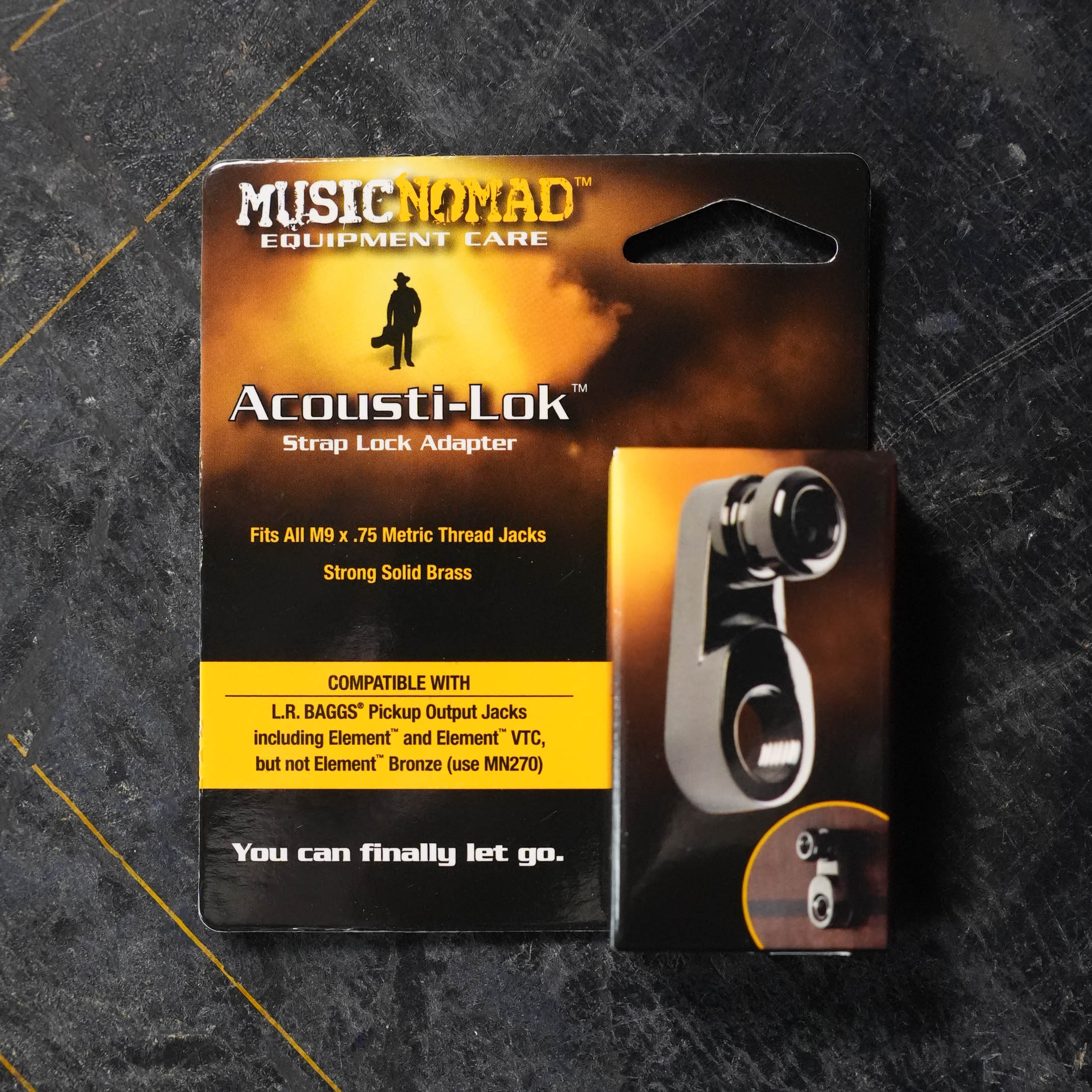A packaged Music Nomad Acousti-Lok Strap Lock Adapter for Metric Output Jacks (MN271) is shown on a dark surface. The packaging highlights LR Baggs pickup compatibility and its construction from solid brass.