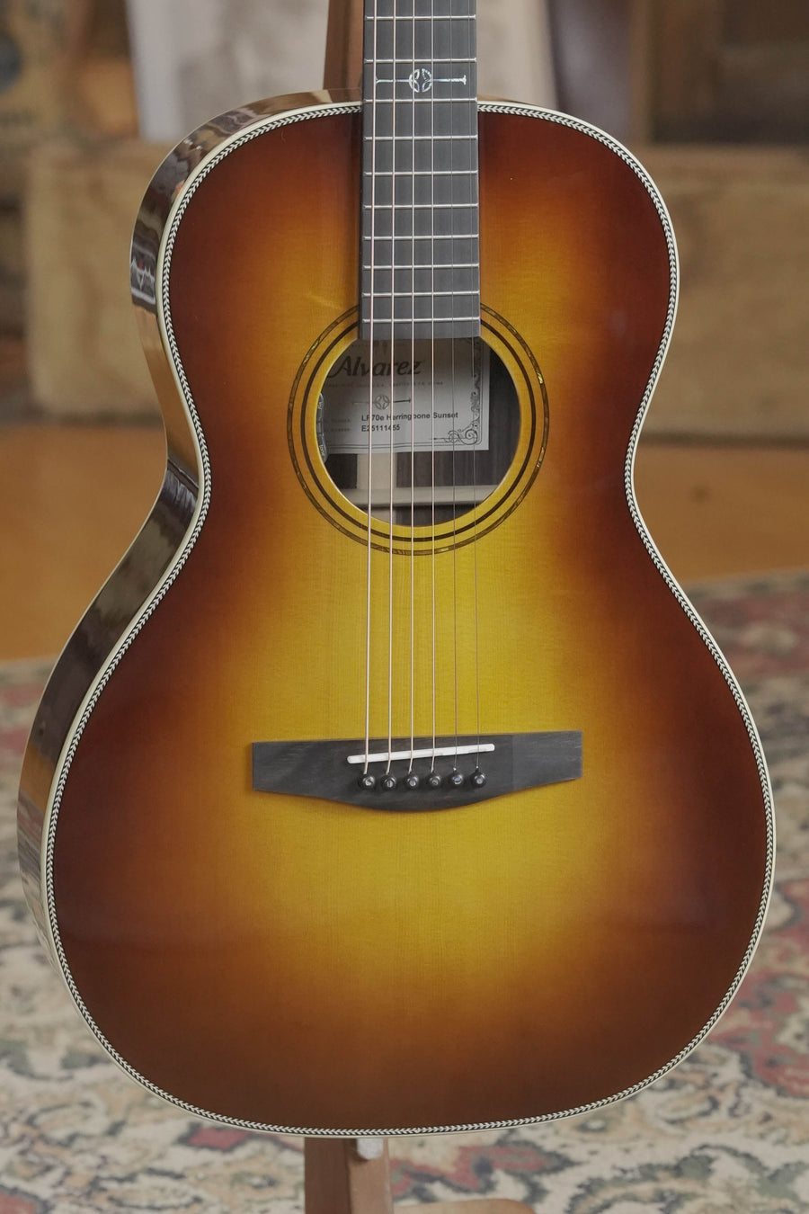 A close-up of the Alvarez Laureate LP70e Herringbone Sunset Parlor Guitar with LR Baggs HiFi Pickup, showcasing its sunburst finish, round soundhole, and six strings, resting on a patterned rug.