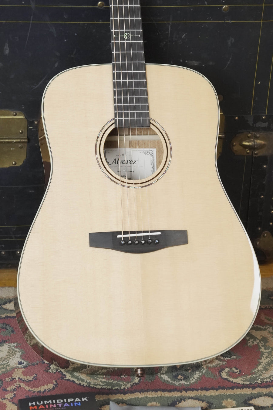 An Alvarez Laureate Series LD60e Sitka/Mahogany Dreadnought Guitar with LR Baggs Hifi Pickup is displayed upright on a patterned rug before a vintage trunk, its label inside the soundhole confirming the Alvarez brand and model.