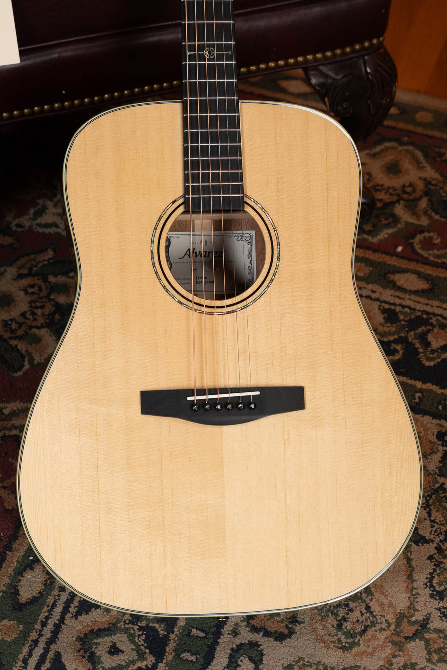 A close-up of the Alvarez Laureate Series LD60e Sitka/Mahogany Dreadnought Guitar w/LR Baggs Hifi Pickup shows its light wood finish, black bridge, and steel strings resting on a patterned rug, with sound hole and fretboard visible.