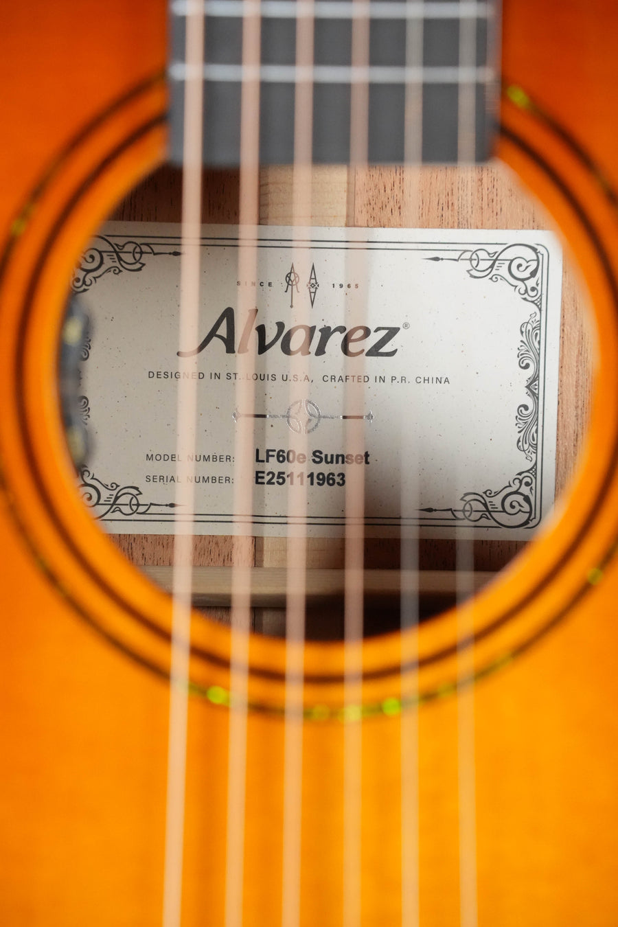 Close-up through the strings of an Alvarez Laureate Series LF60e Sunset Sitka/Mahogany OM Guitar, showing the inner label with model and serial numbers. The glossy orange finish enhances its acoustic resonance. Features LR Baggs Hifi Pickup.