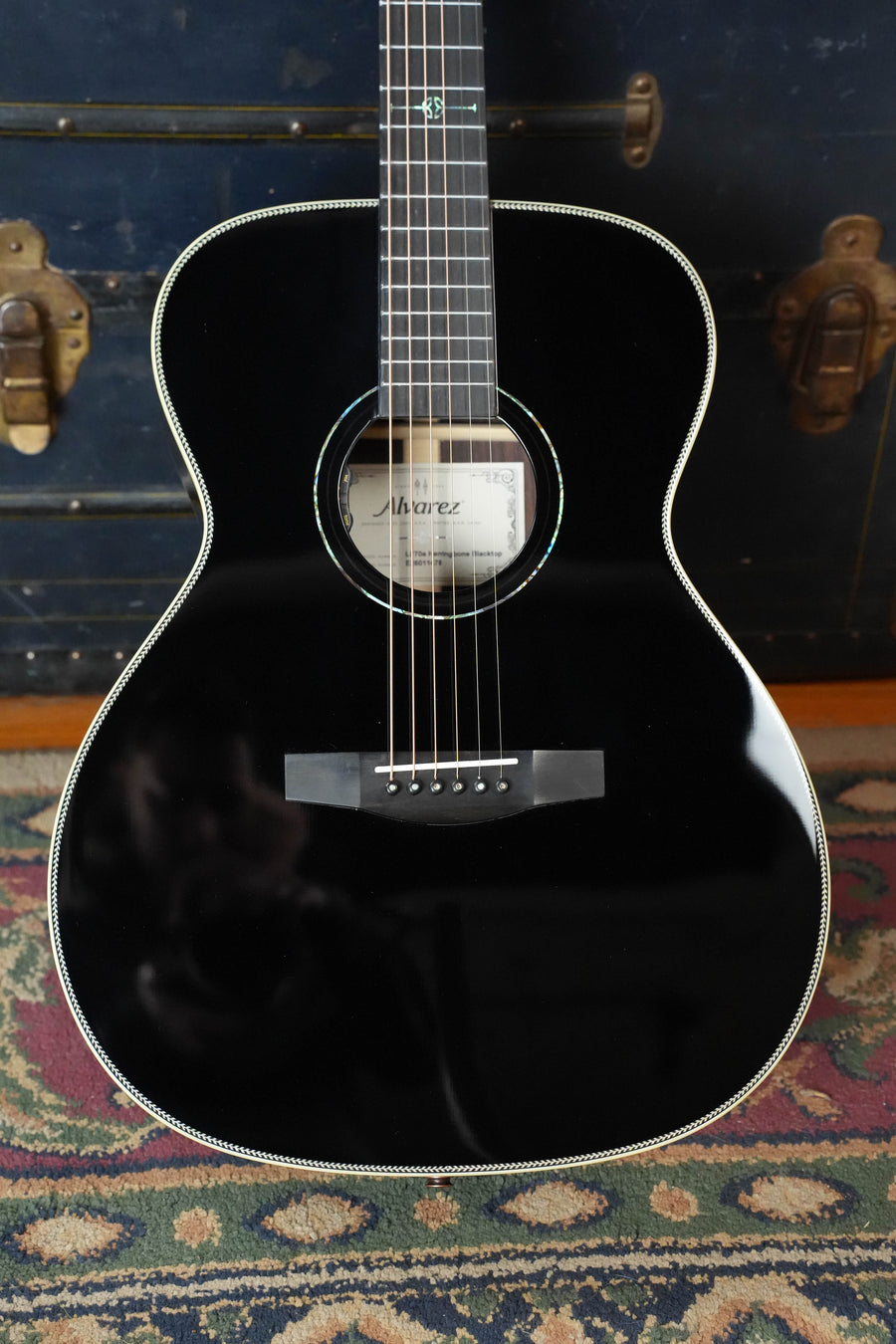 An Alvarez Laureate Series LF70e Herringbone Blacktop Sitka/Rosewood Sunset OM Guitar w/LR Baggs Hifi Pickup (#1478) stands upright on a patterned rug, its glossy finish reflecting surroundings, with a vintage brass-trimmed trunk behind.