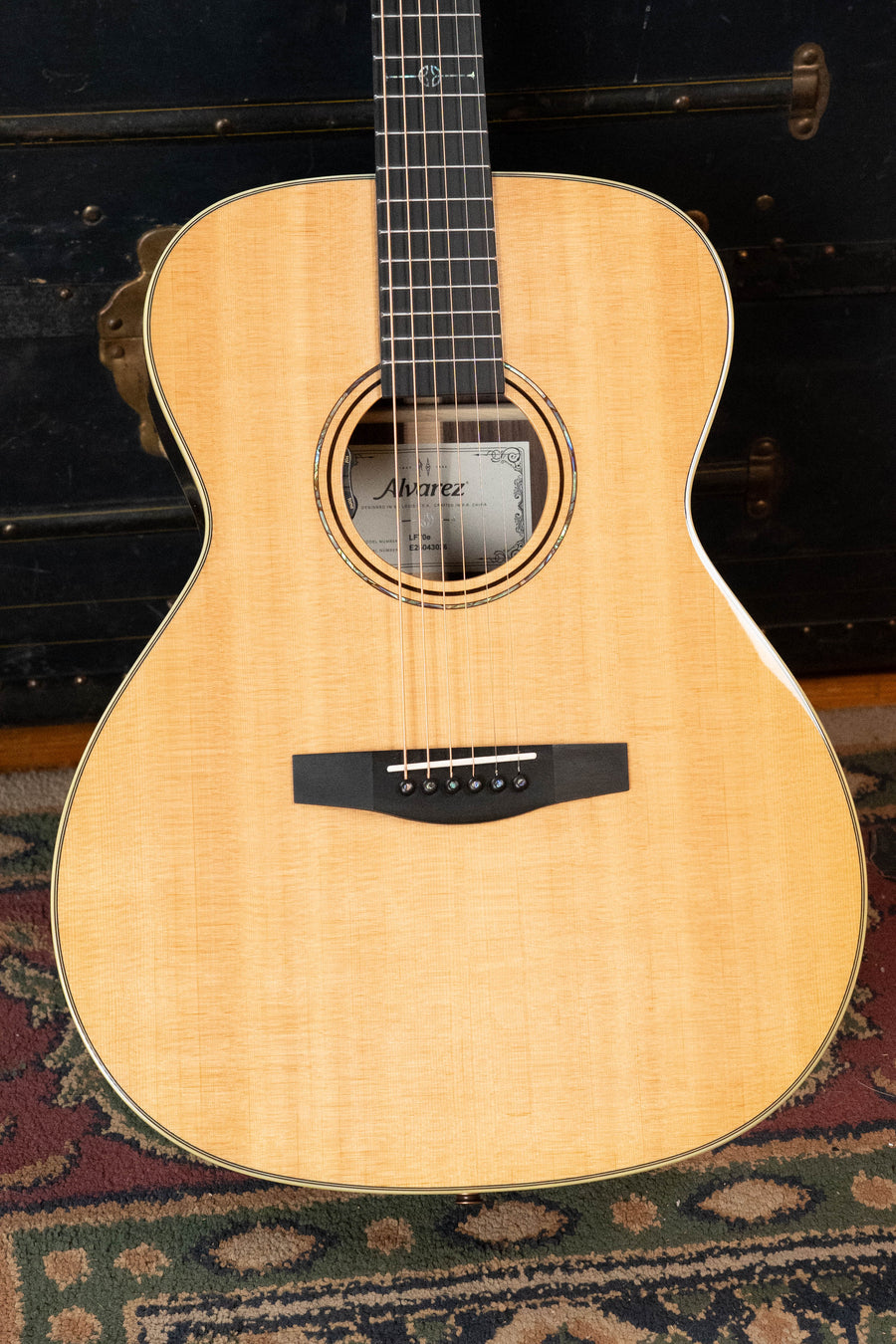 A close-up of the Alvarez Laureate Series LF70e Herringbone Sitka/Rosewood OM w/LR Baggs Hifi Pickup #3036 shows its solid Sitka spruce top and six strings, set against a patterned rug with a dark trunk in the background.