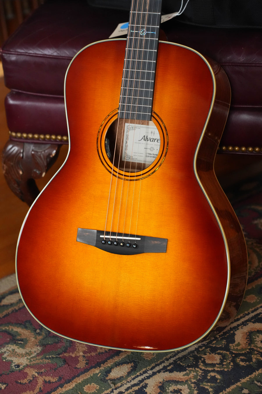 An Alvarez Laureate Series LT60e Sunset Sitka/Mahogany OOO Guitar w/LR Baggs Hifi Pickup (#1561) rests on a patterned rug by a dark leather chair, its label reading Alvarez inside the sound hole.