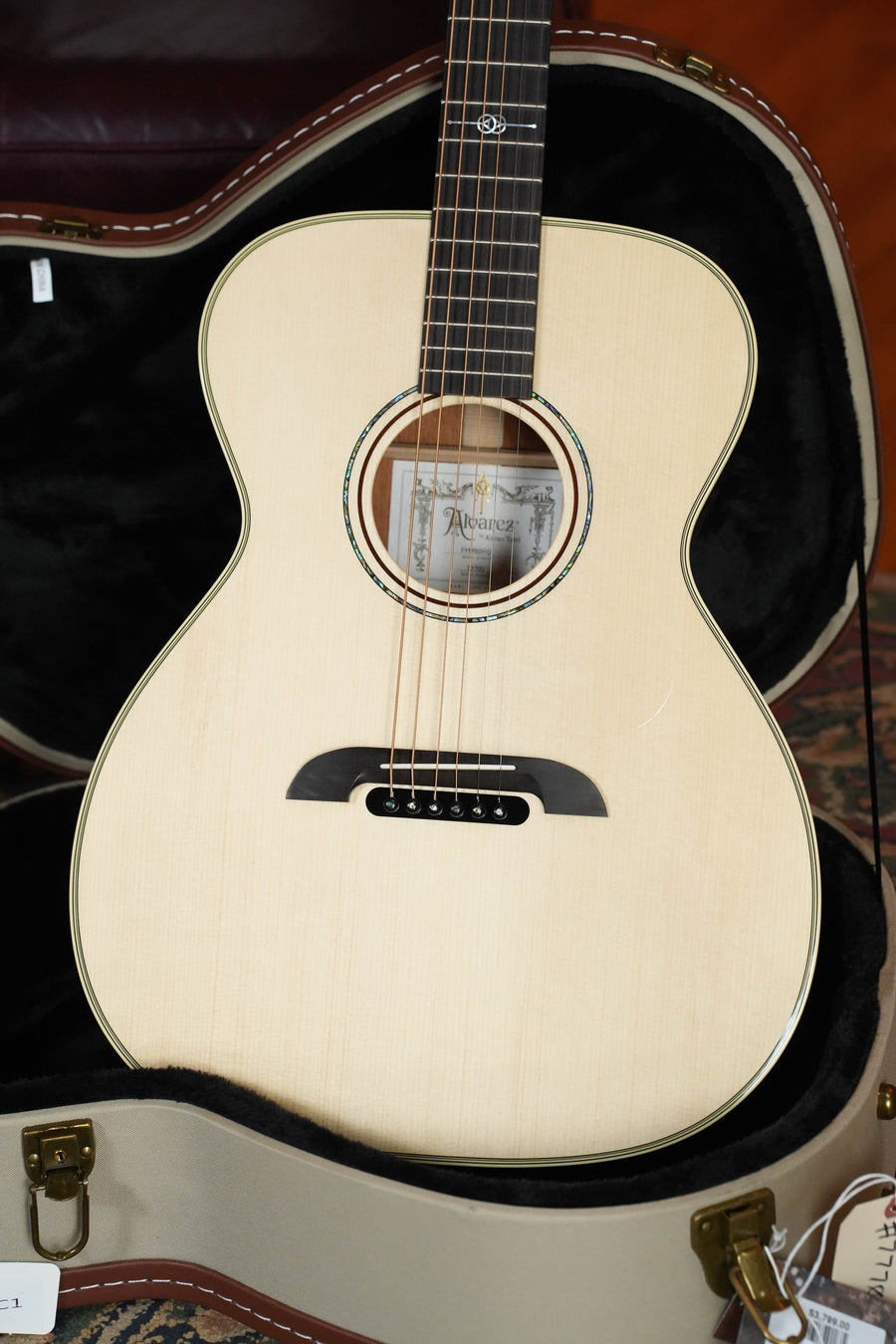 An Alvarez Yairi Honduran Series FYM60HD OM Acoustic Guitar with an Adirondack Spruce top rests in an open, padded case featuring a black interior and tan exterior. A paper tag hangs from the case latch.