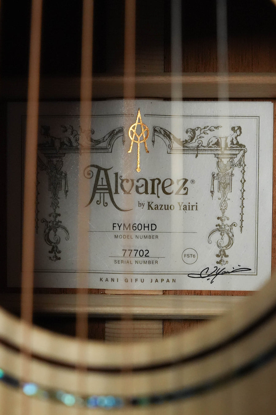 Close-up of an Alvarez Yairi Honduran Series FYM60HD Adirondack/Vintage Honduran Mahogany OM Acoustic Guitar #7702 label inside the soundhole, showing the brand and model with decorative borders and guitar strings in the foreground.