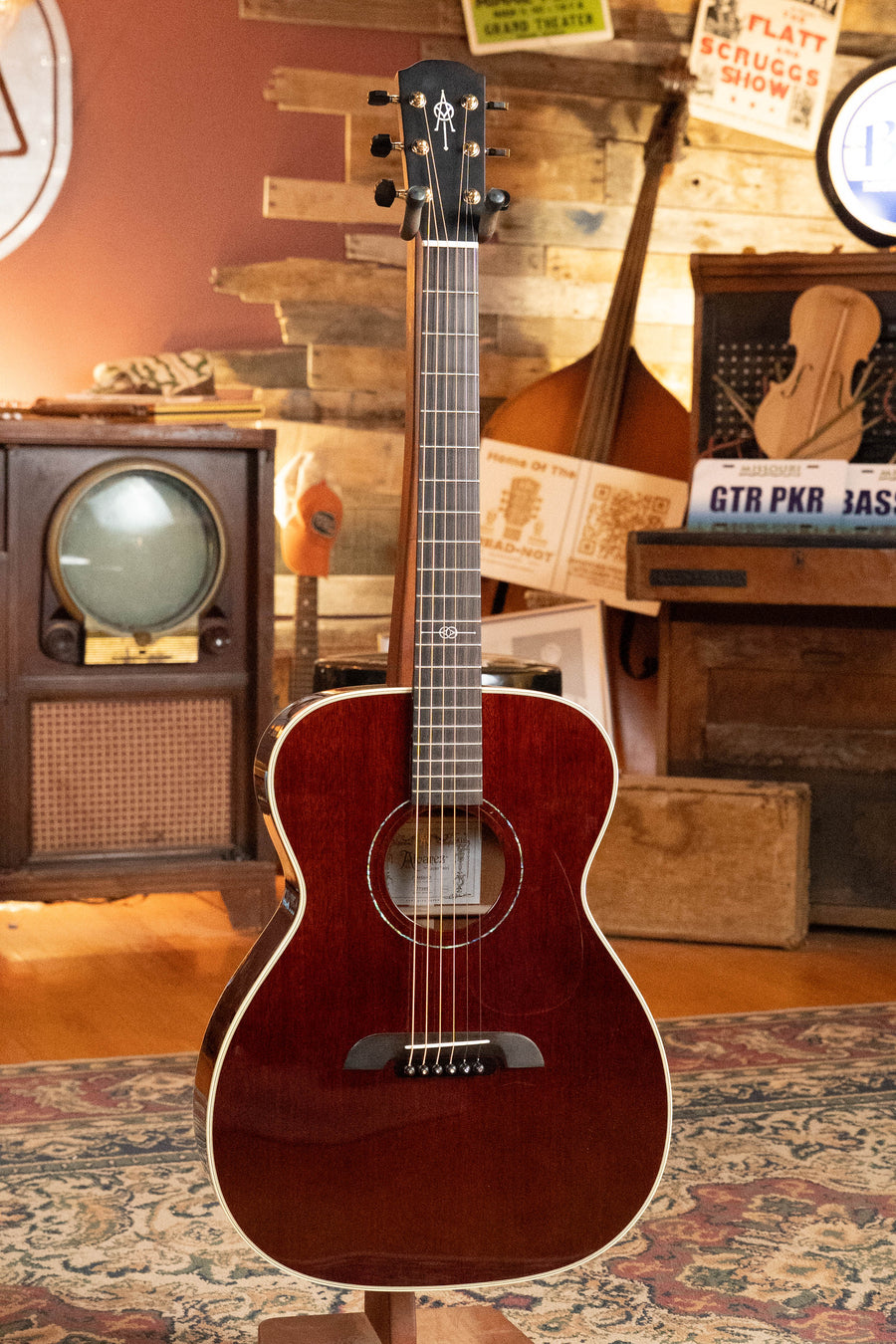 An Alvarez Yairi Honduran Series FYM66HD All Vintage Mahogany OM Acoustic Guitar #7595 by Alvarez stands upright on a patterned rug in a cozy room featuring a vintage TV, upright bass, wood-paneled wall, and music-themed decor.