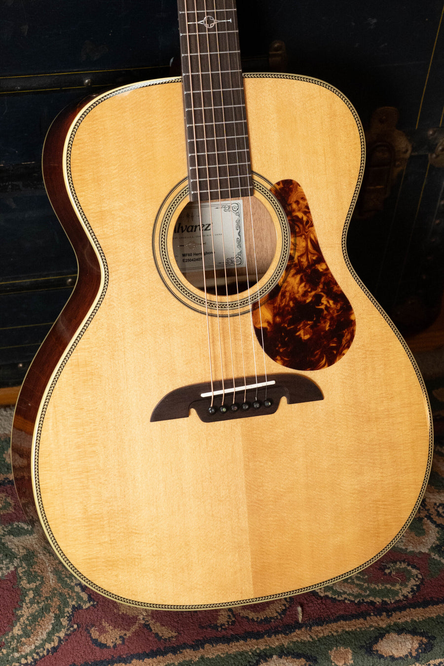 An Alvarez Masterworks Series MF60 Herringbone Sitka/African Mahogany Orchestra Model acoustic guitar, featuring a light Sitka spruce top, dark African mahogany sides, and a tortoiseshell-style pickguard, rests upright on a patterned rug. The Alvarez brand is visible on the label inside the sound hole.