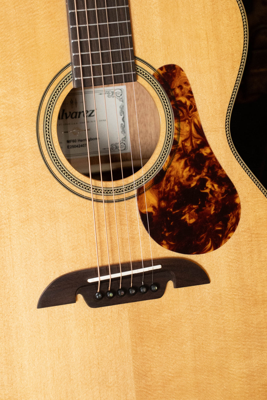 Close-up of an Alvarez Masterworks Series MF60 Herringbone Sitka/African Mahogany Orchestra Model Acoustic Guitar (#2407) by Alvarez, highlighting the strings, sound hole with interior label, decorative herringbone rosette, dark tortoiseshell-style pickguard, and wooden bridge on a light-colored solid Sitka spruce top.