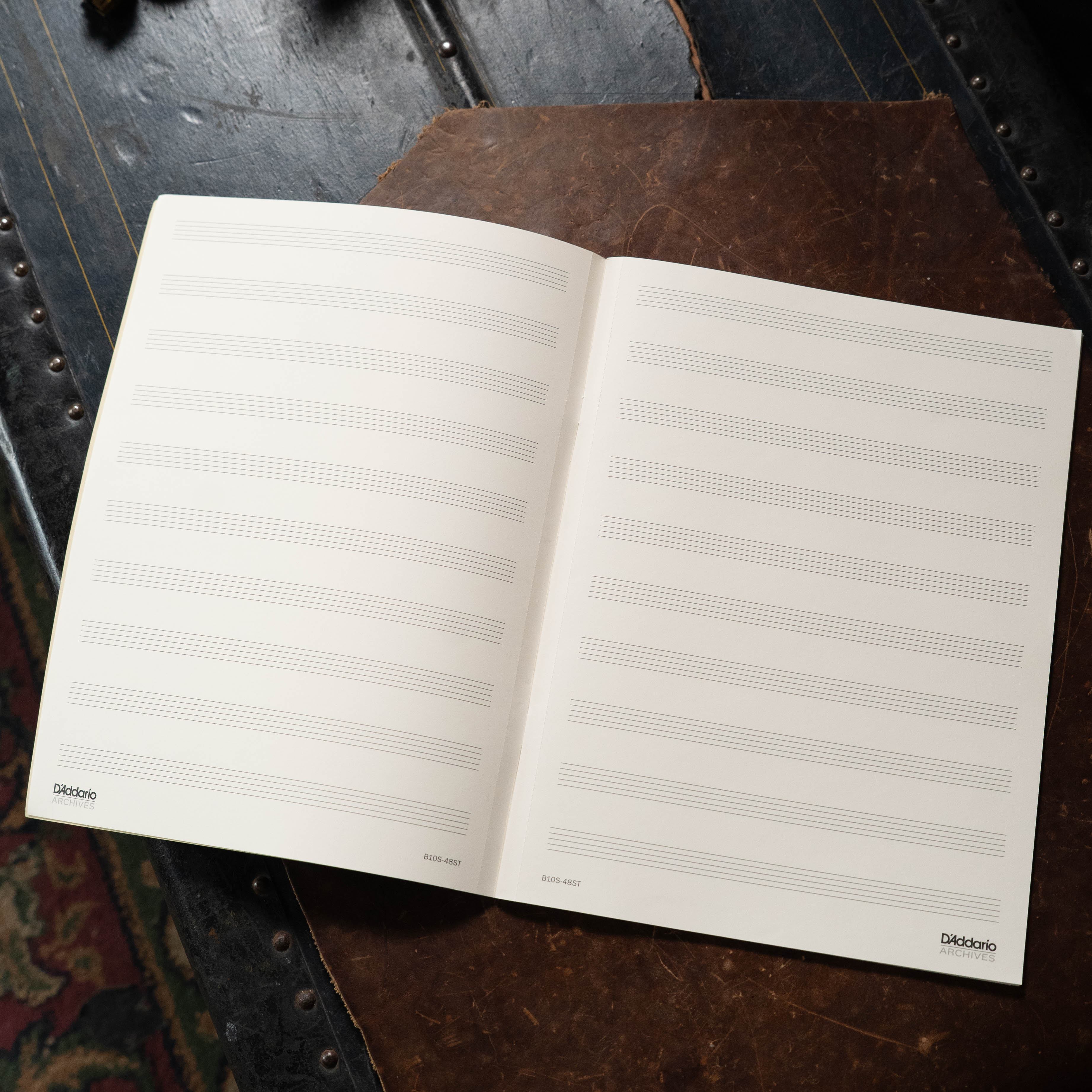 An open Archives 48 pg 10 Stave book by The Acoustic Shoppe rests on a dark wooden surface, displaying two 9x12 manuscript pages with 10 staves each. The notebook's cover is partially visible beneath the open pages.