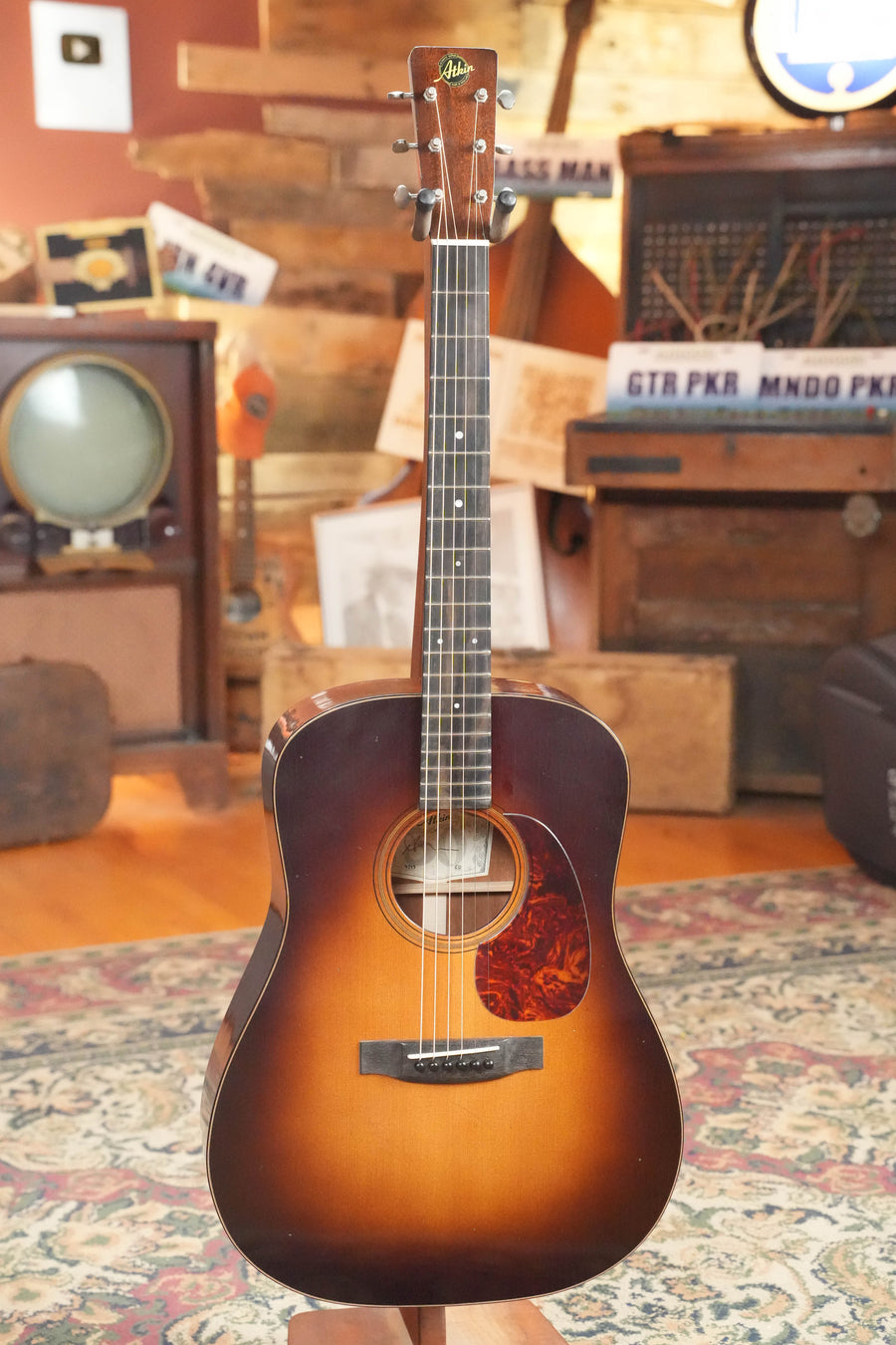 An Atkin Guitars Essential D 45MM Sitka/Mahogany Dreadnought Acoustic Guitar (#4399) stands upright in a cozy room, surrounded by a patterned rug, old TV, wooden furniture, and retro decor.