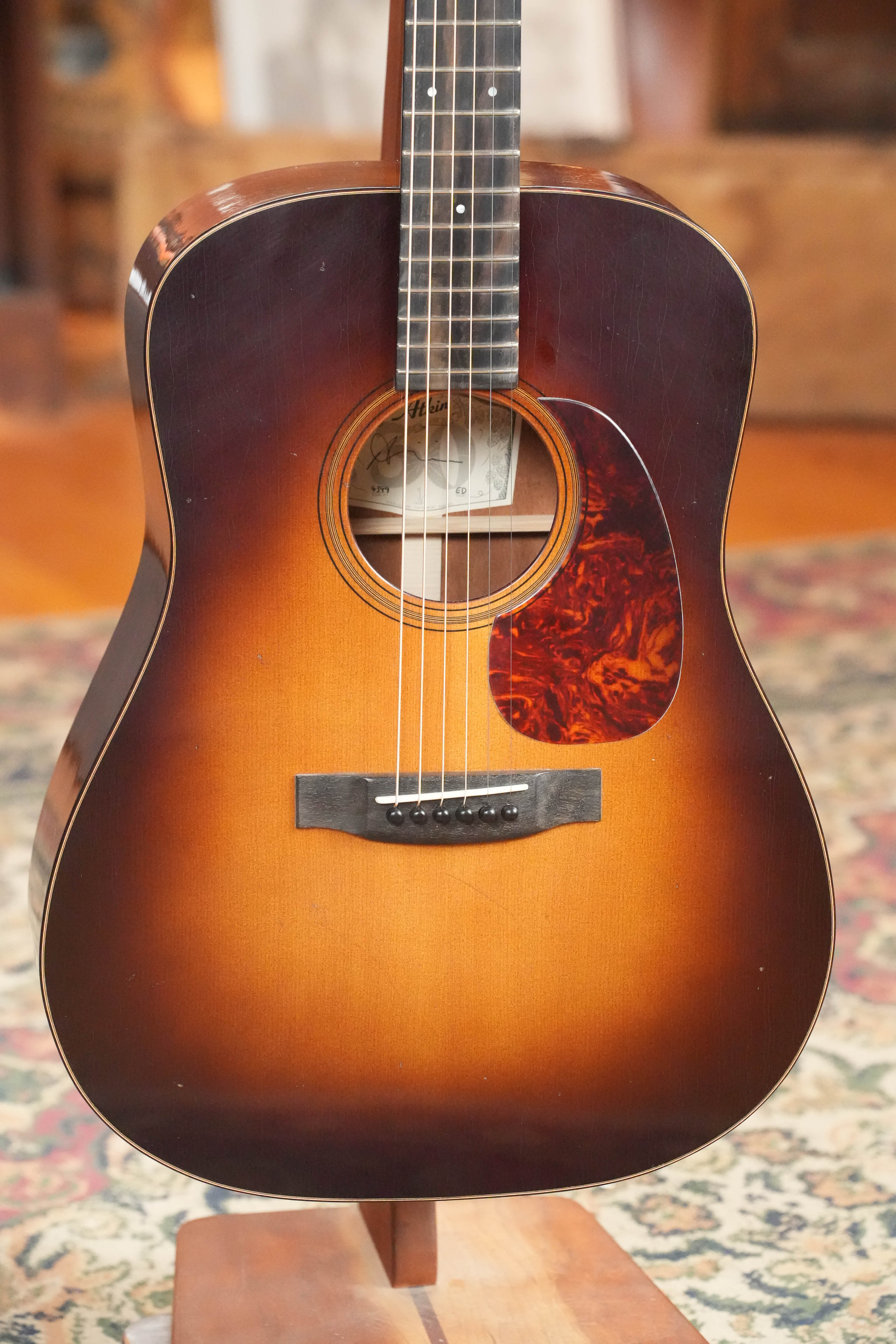 An Atkin Guitars Essential D 45MM Sitka/Mahogany Dreadnought Acoustic Guitar (#4399) with a tortoiseshell-patterned pickguard rests on a wooden stand atop a patterned rug in a warmly lit room.