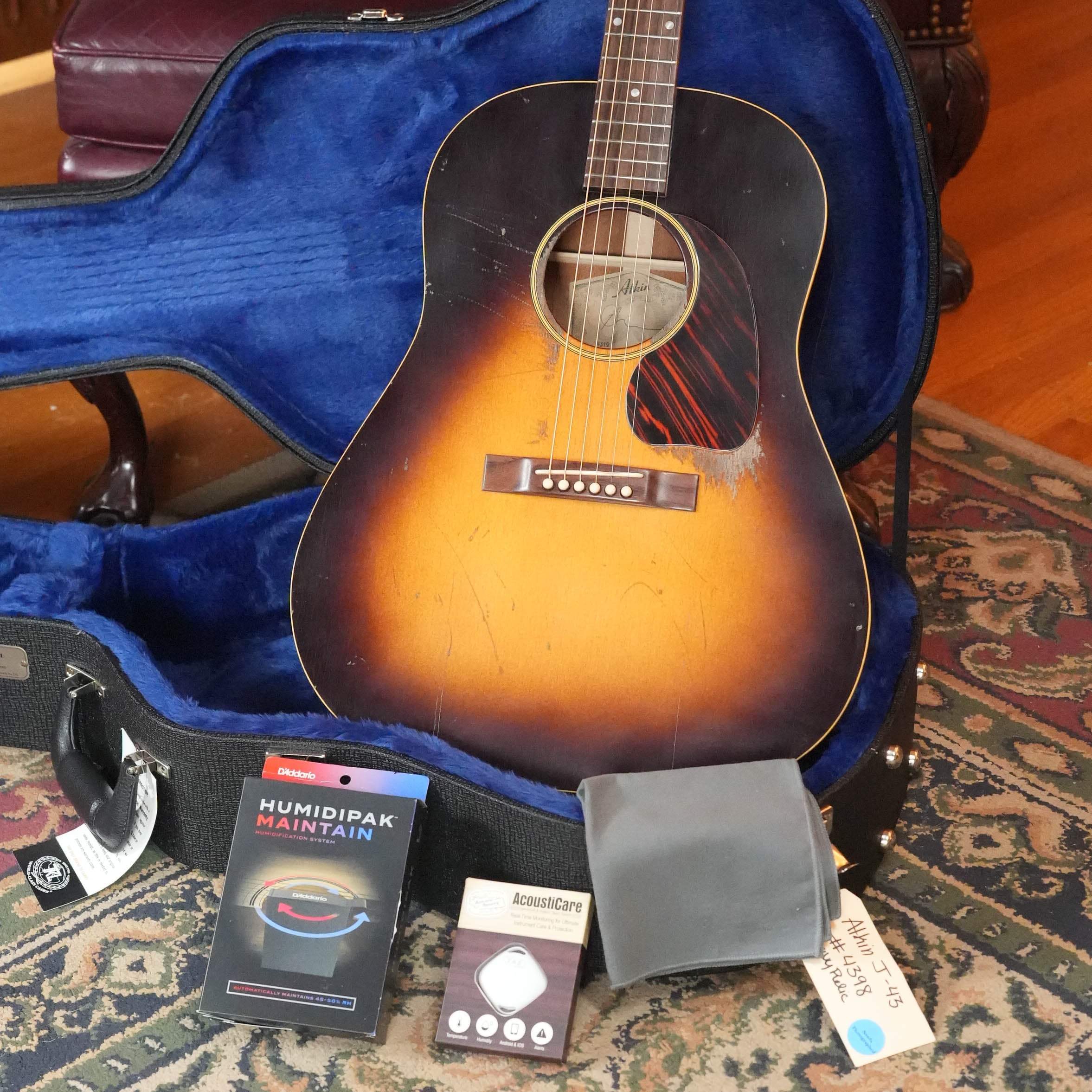 An Atkin J43 45MM Baked Sitka/Mahogany Heavy Aged Slope Shoulder Dreadnought Acoustic Guitar by Atkin Guitars rests in an open blue-lined case on a patterned rug, with a Humidipak Maintain kit, gray pouch, AcousCare box, and handwritten “1955 J-45” tag nearby.