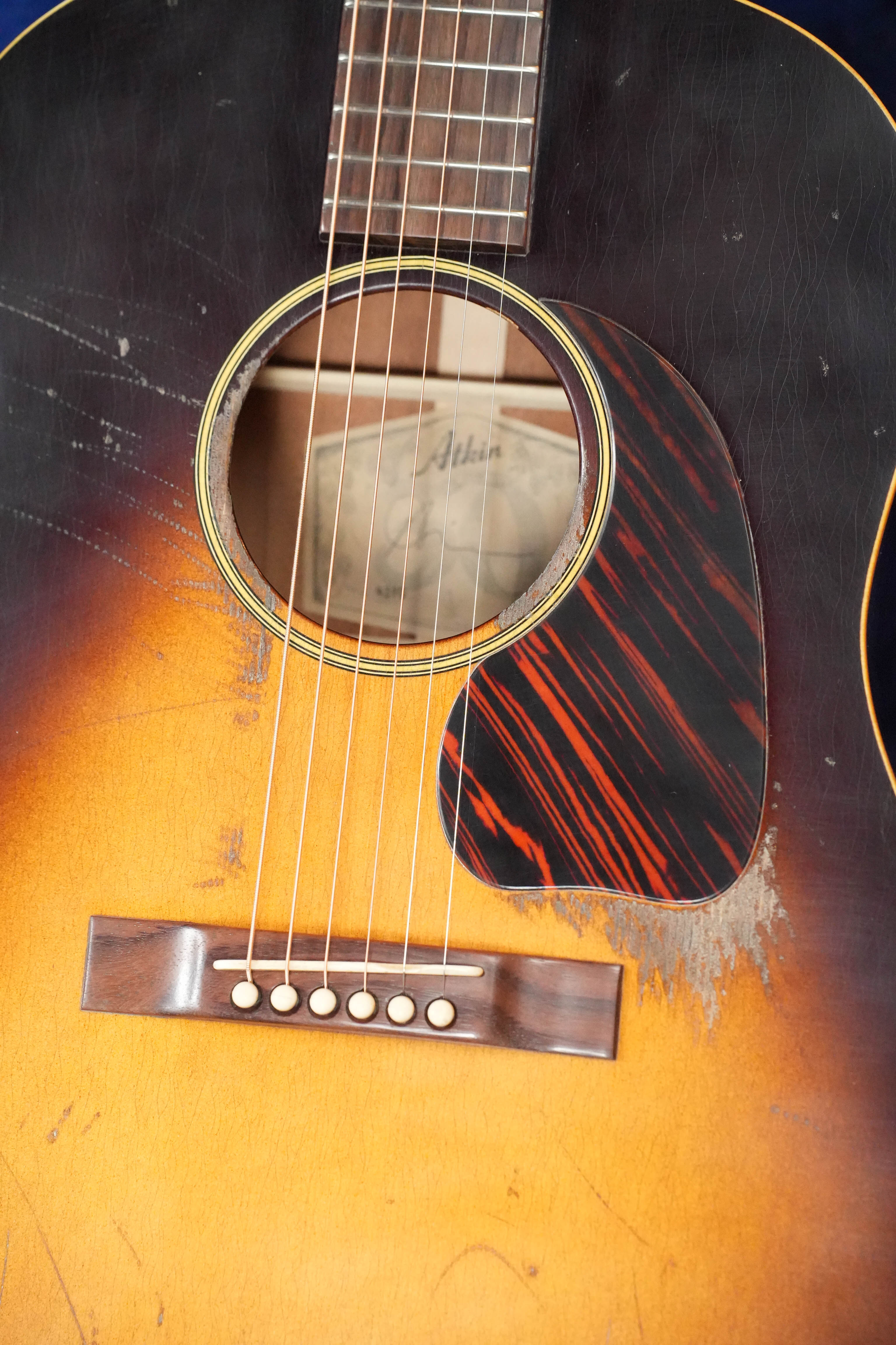 A close-up of the Atkin J43 45MM Baked Sitka/Mahogany Heavy Aged Slope Shoulder Dreadnought Acoustic Guitar by Atkin Guitars shows its sunburst finish, tortoiseshell pickguard, visible pick scratches, six strings, and a paper label inside the soundhole.