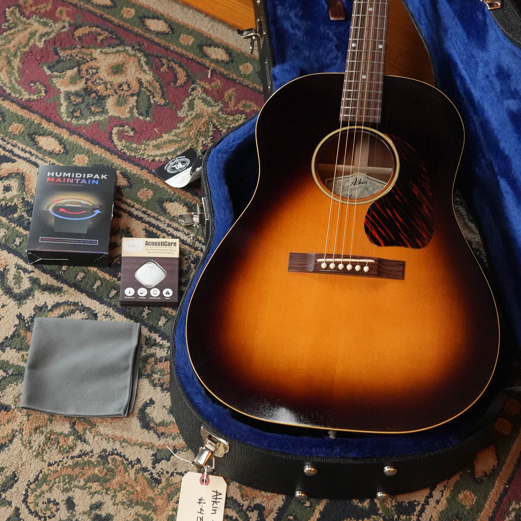 The Atkin Guitars J43 Baked Sitka/Mahogany Heavy Aged Slope Shoulder Dreadnought Acoustic Guitar #4397 rests in a blue-lined case on a patterned rug, with gray cloth, humidipak, accessory, and black device arranged nearby.