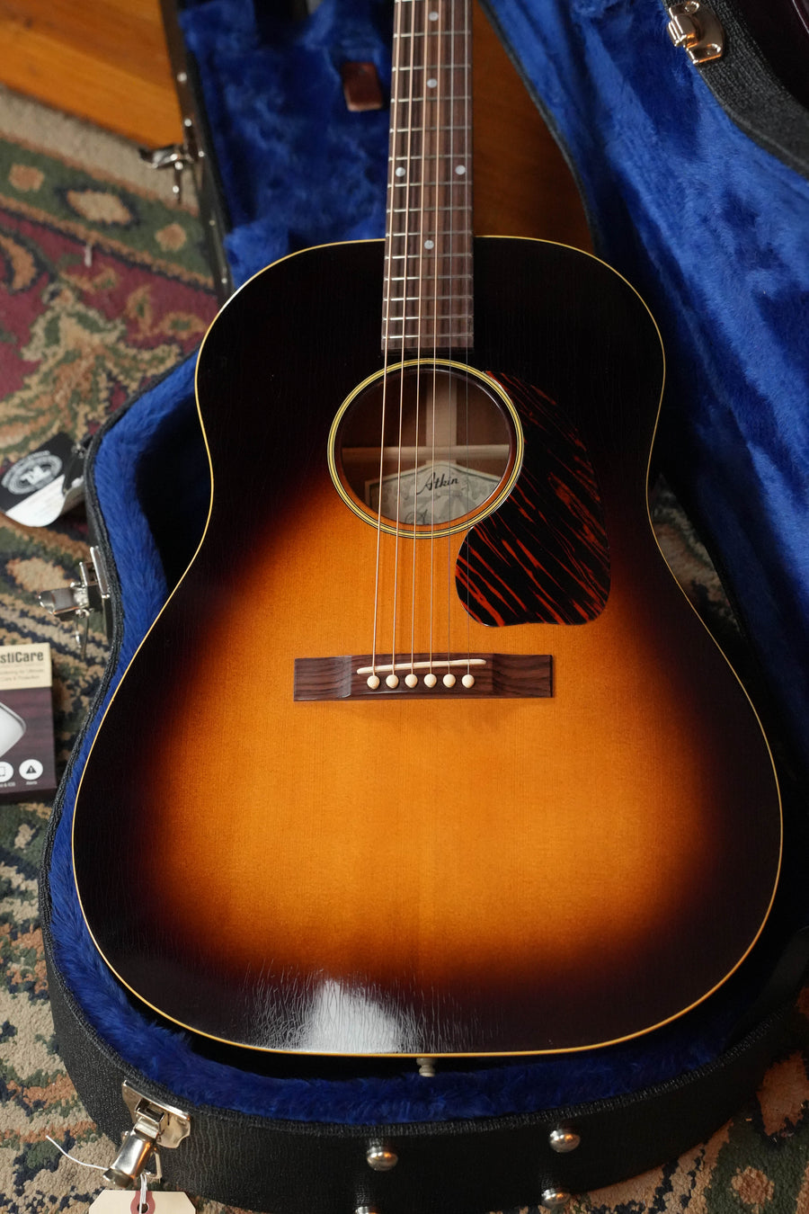 An Atkin Guitars J43 Baked Sitka/Mahogany Heavy Aged Slope Shoulder Dreadnought Acoustic Guitar (#4397) with a sunburst gloss finish rests in a blue plush-lined hard case, with vintage accessories and a patterned rug nearby.