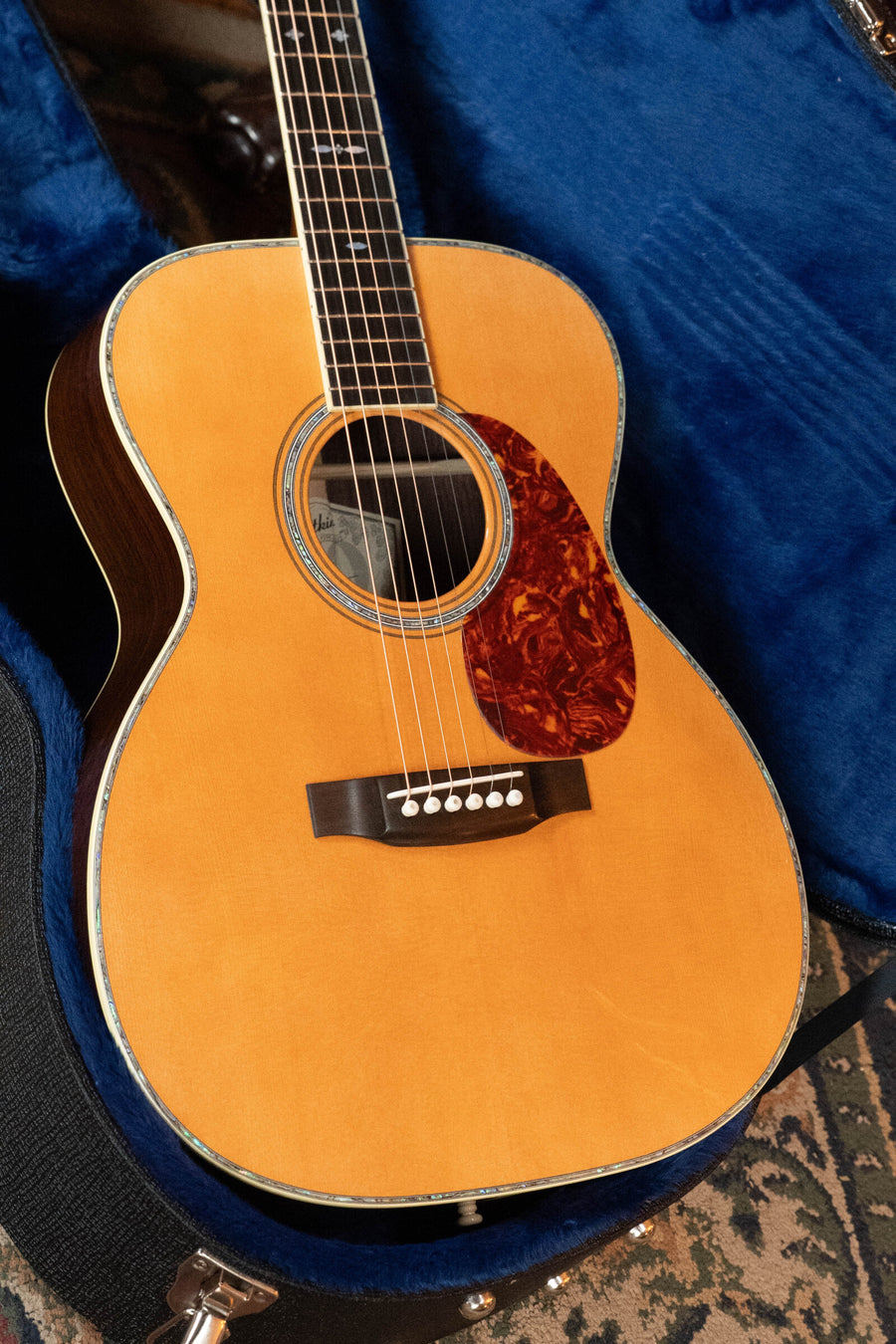 An Atkin Guitars 00037 DLX Sitka/East Indian Rosewood OOO Acoustic Guitar #3895, featuring a natural wood and aged nitrocellulose finish, rests in a blue-lined hard case, showcasing a tortoiseshell pickguard and decorative binding inspired by classic pre-war triple-0 designs.