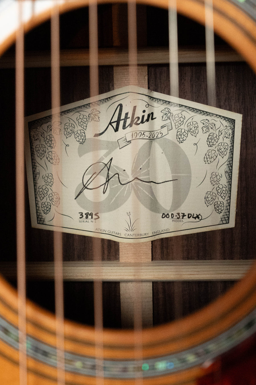 Close-up of a guitar’s soundhole showing a label inside. The label reads Atkin 1995-2023 and displays a signature, serial number, and decorative hops illustrations. This Atkin Guitars 00037 DLX Sitka/East Indian Rosewood OOO Acoustic Guitar #3895 features an aged nitrocellulose finish, capturing vintage character inspired by classic pre-war triple-0 guitars.