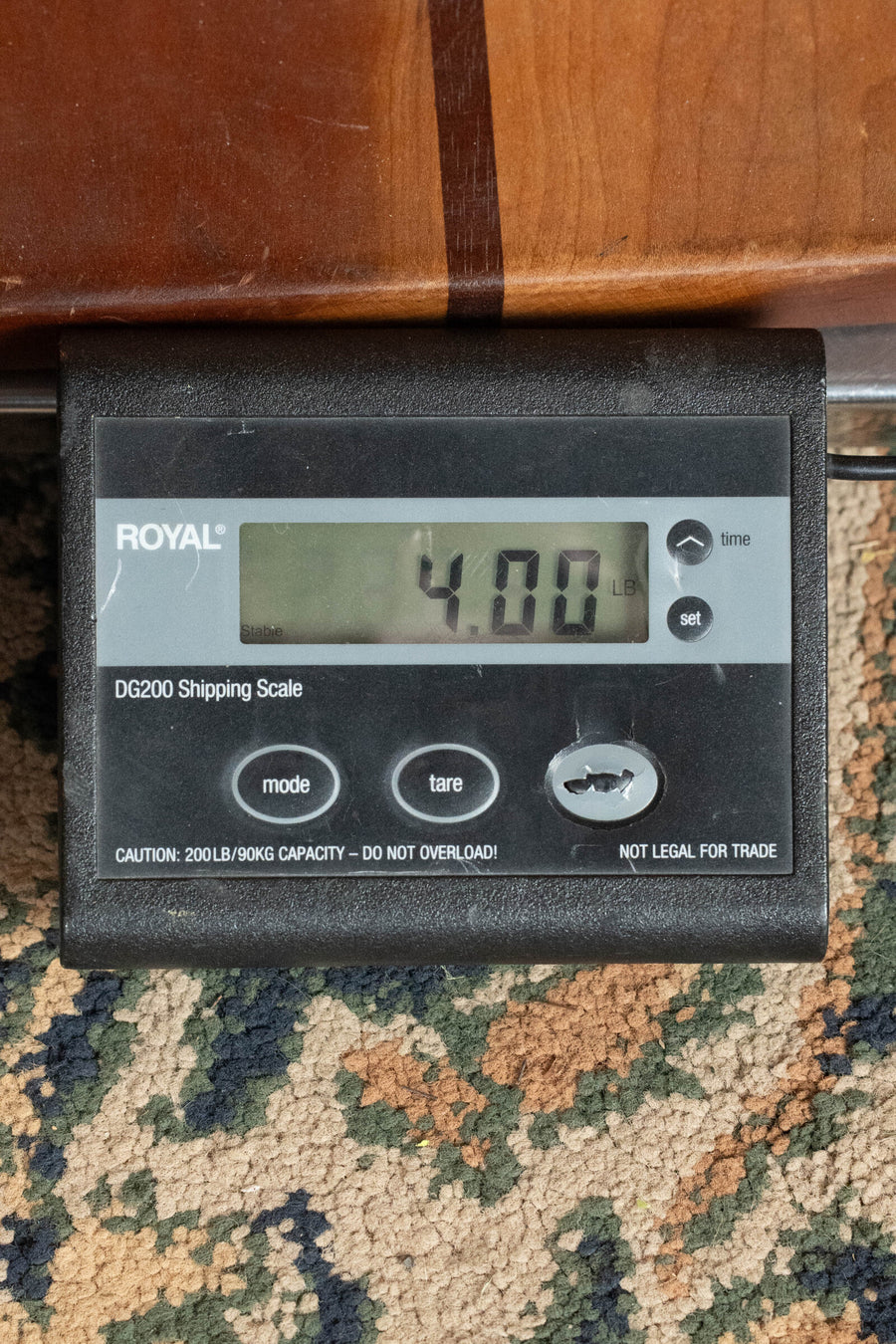 A digital shipping scale displays a weight of 4.00 lbs, calling to mind the precision found in the Atkin Guitars 00037 DLX Sitka/East Indian Rosewood OOO Acoustic Guitar #3895. The scale sits on a patterned carpet with part of a wooden surface above it, and its screen shows buttons labeled mode, tare, and set.