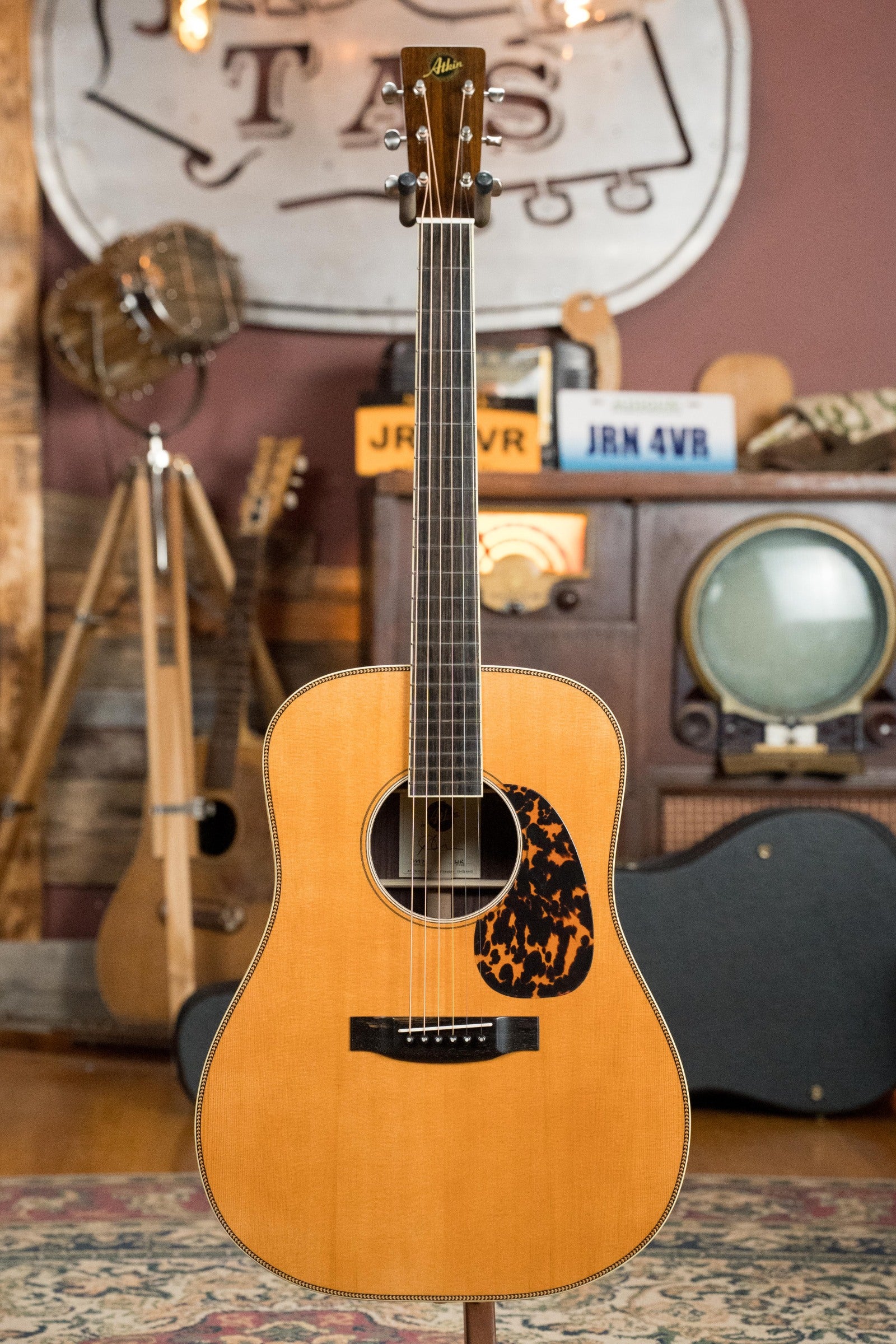 An Atkin Guitars White Rice Baked Sitka/Rosewood Dreadnought Acoustic Guitar (#3497) with a natural finish and tortoiseshell pickguard stands on a stand, exuding classic charm among vintage radios, instruments, and decor in the background.