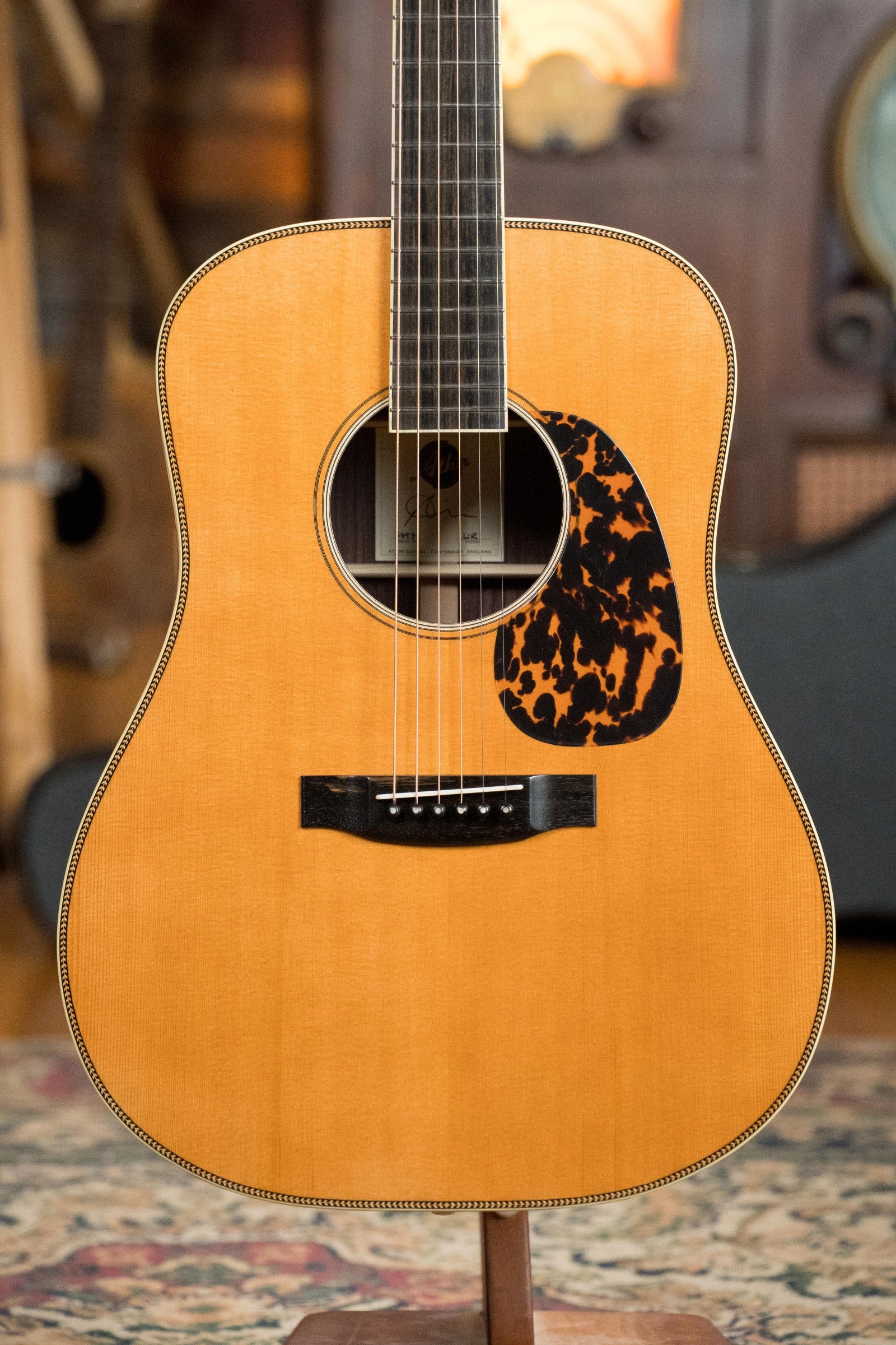 A close-up of the Atkin Guitars White Rice Baked Sitka/Rosewood Dreadnought Acoustic Guitar #3497 with a light wood body and tortoiseshell pickguard, standing on a rug against a blurred background of musical instruments.