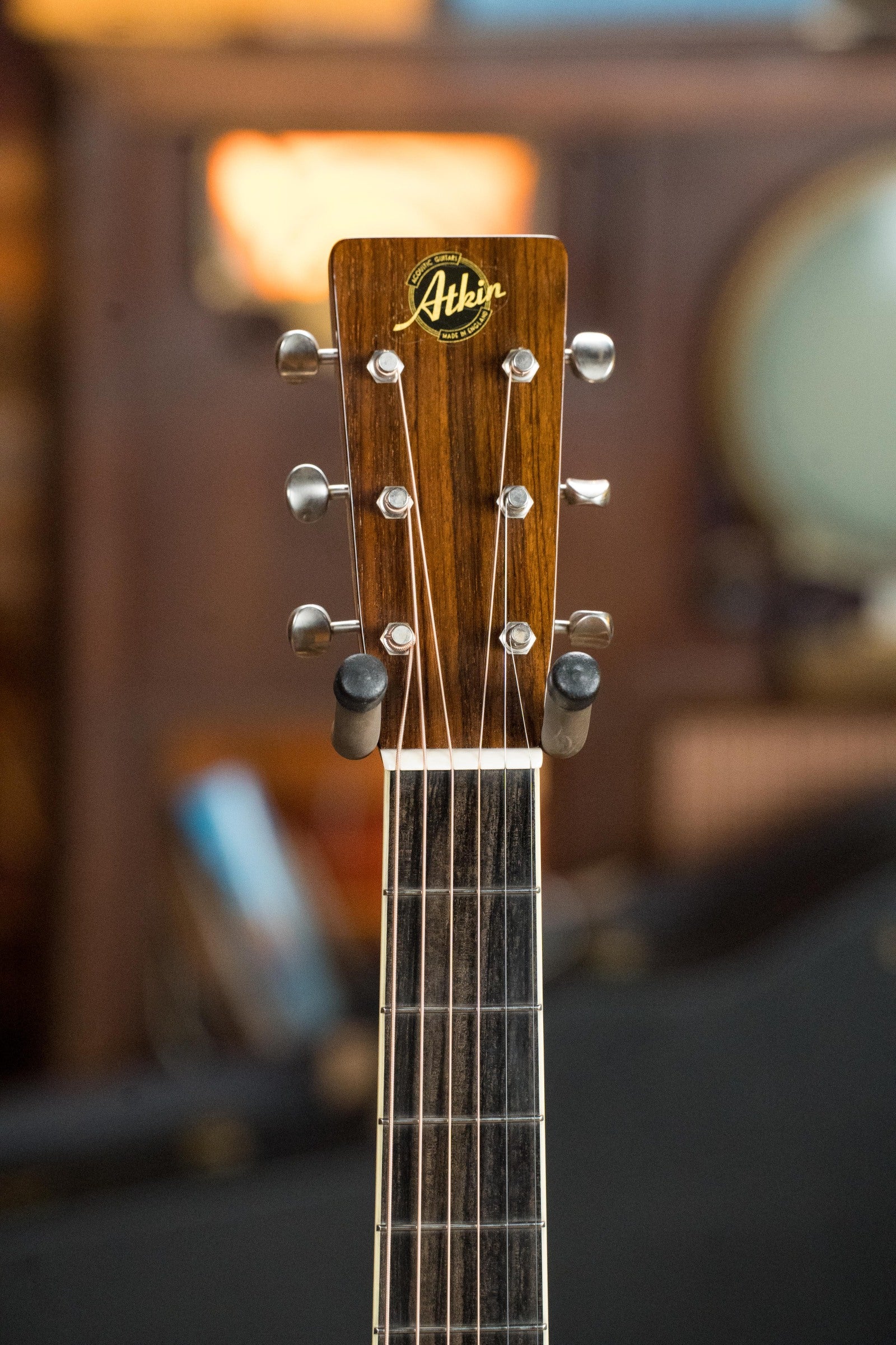 Close-up of an Atkin Guitars White Rice Baked Sitka/Rosewood Dreadnought Acoustic Guitar #3497 headstock, featuring the Atkin logo, six tuning pegs, and metal strings with a blurred indoor background.