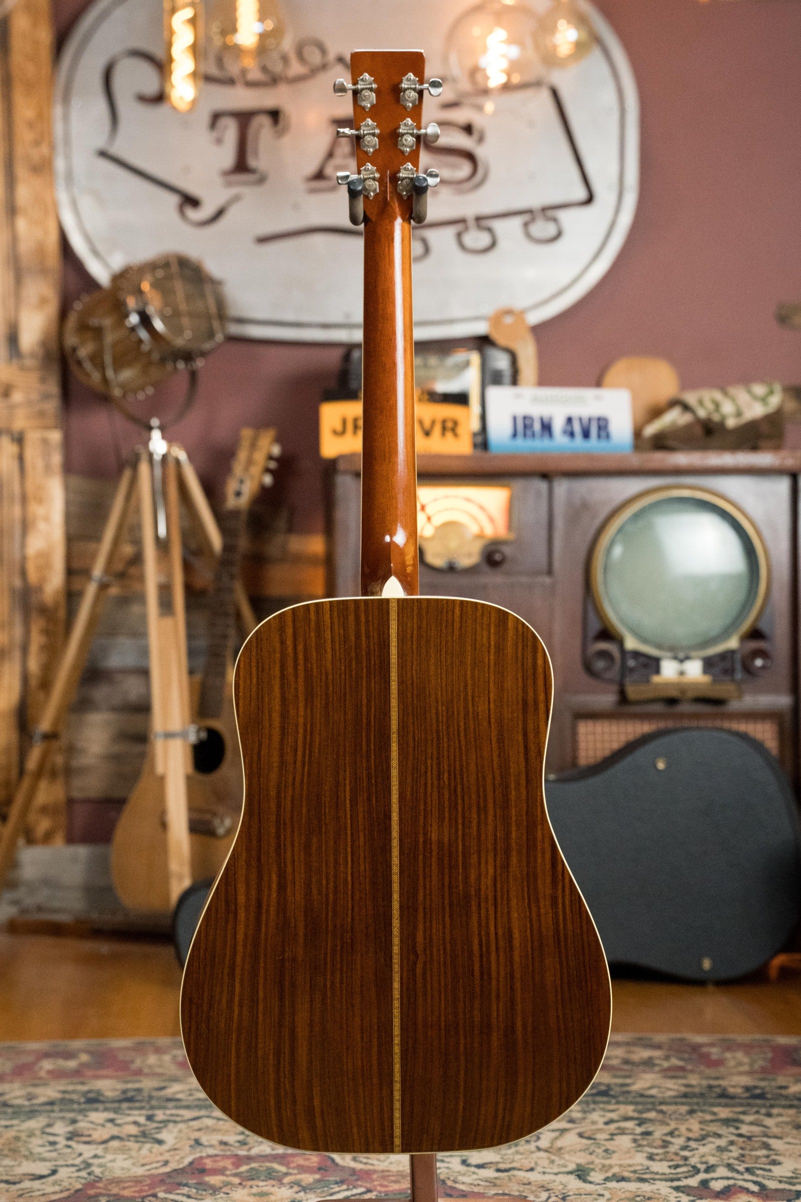 An Atkin Guitars White Rice Baked Sitka/Rosewood Dreadnought Acoustic Guitar (#3497) with a glossy finish and light center stripe is shown from the back, upright in a room decorated with vintage amps and a retro TV.
