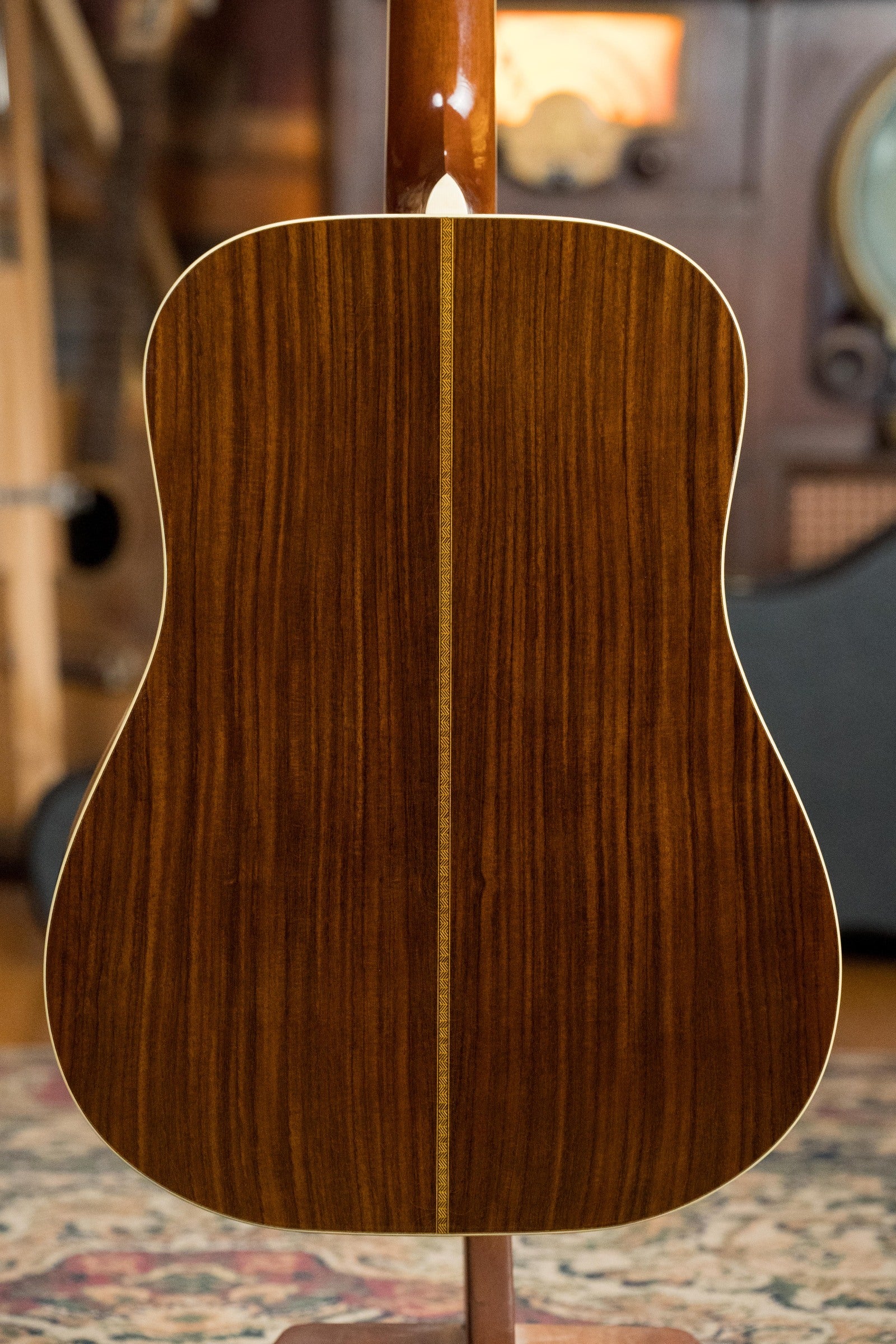 The back of an Atkin Guitars White Rice Baked Sitka/Rosewood Dreadnought Acoustic Guitar #3497, featuring Indian rosewood with a light wood inlay, photographed indoors on a rug with blurred instruments in the background.