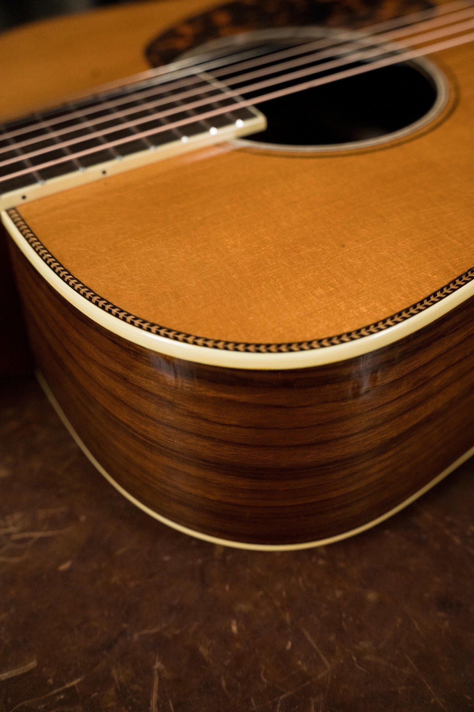 Close-up of an Atkin Guitars White Rice Baked Sitka/Rosewood Dreadnought Acoustic Guitar #3497, showing Indian rosewood grain, decorative binding, strings, and sound hole as it rests on a brown surface.