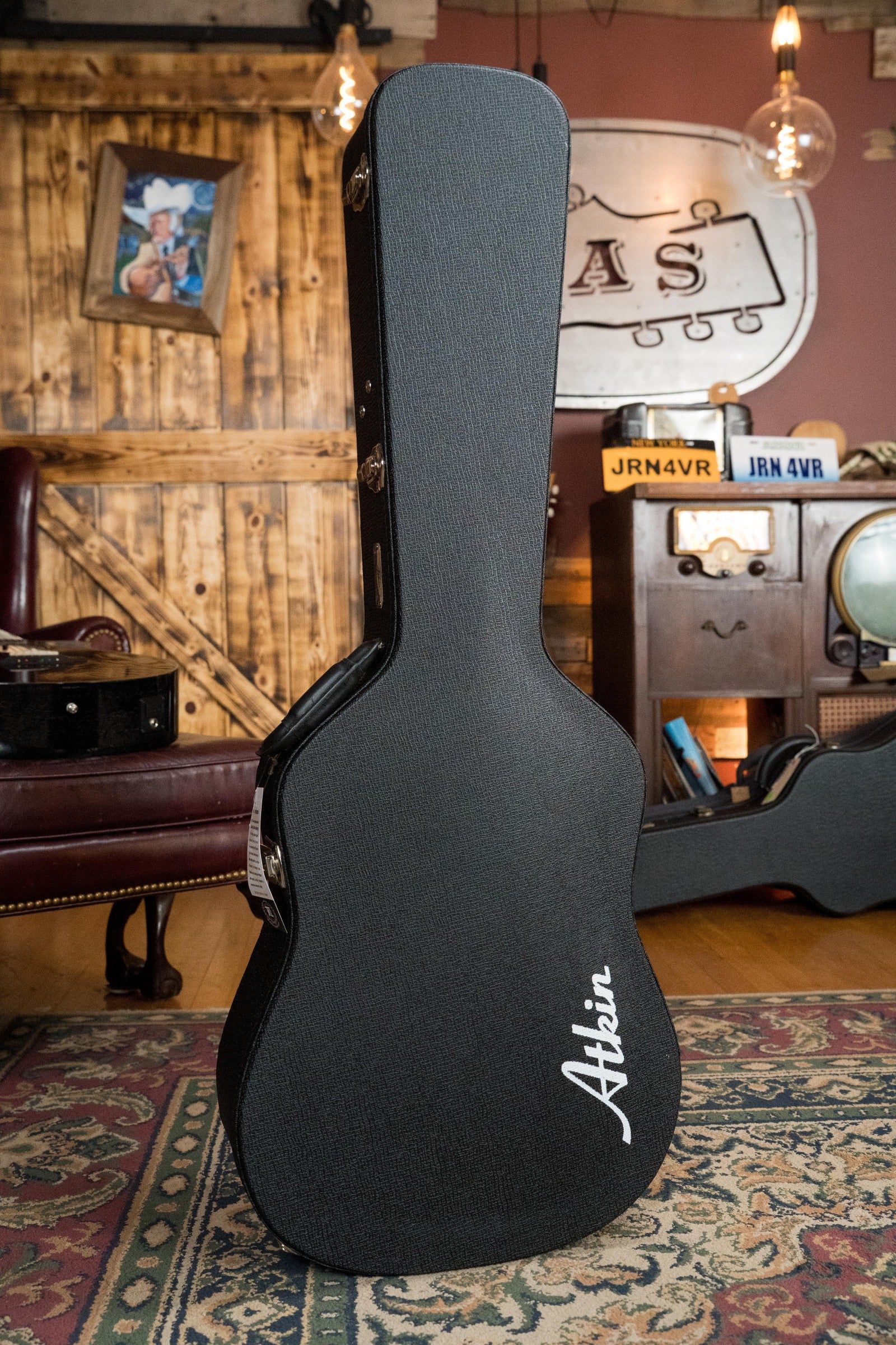 A black guitar case with Atkin Guitars branding stands upright in a cozy, rustic room—an ideal home for the Atkin White Rice Baked Sitka/Rosewood Dreadnought Acoustic Guitar #3497 or another vintage-style instrument.