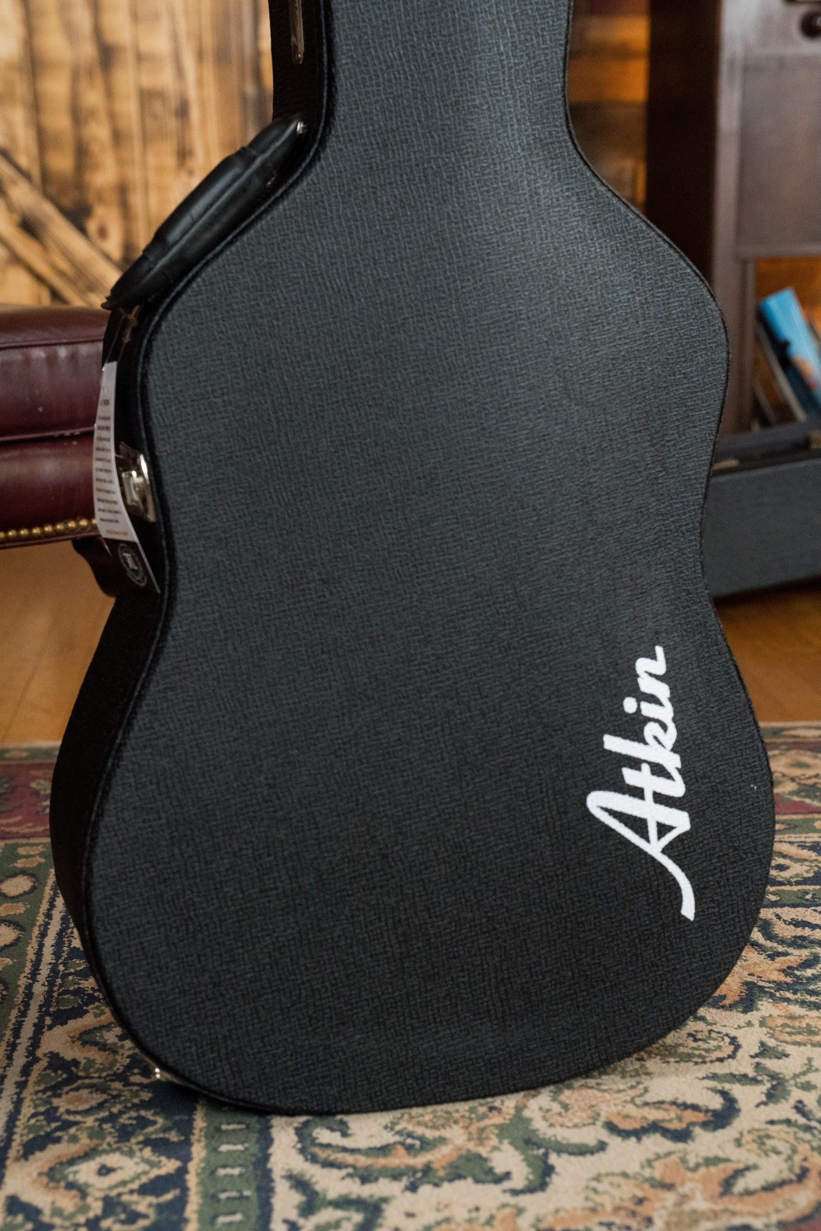 A black Atkin Guitars case with white script rests on a patterned rug, suggesting the Atkin White Rice Baked Sitka/Rosewood Dreadnought Acoustic Guitar #3497 inside, with wooden furniture and part of a room in view.
