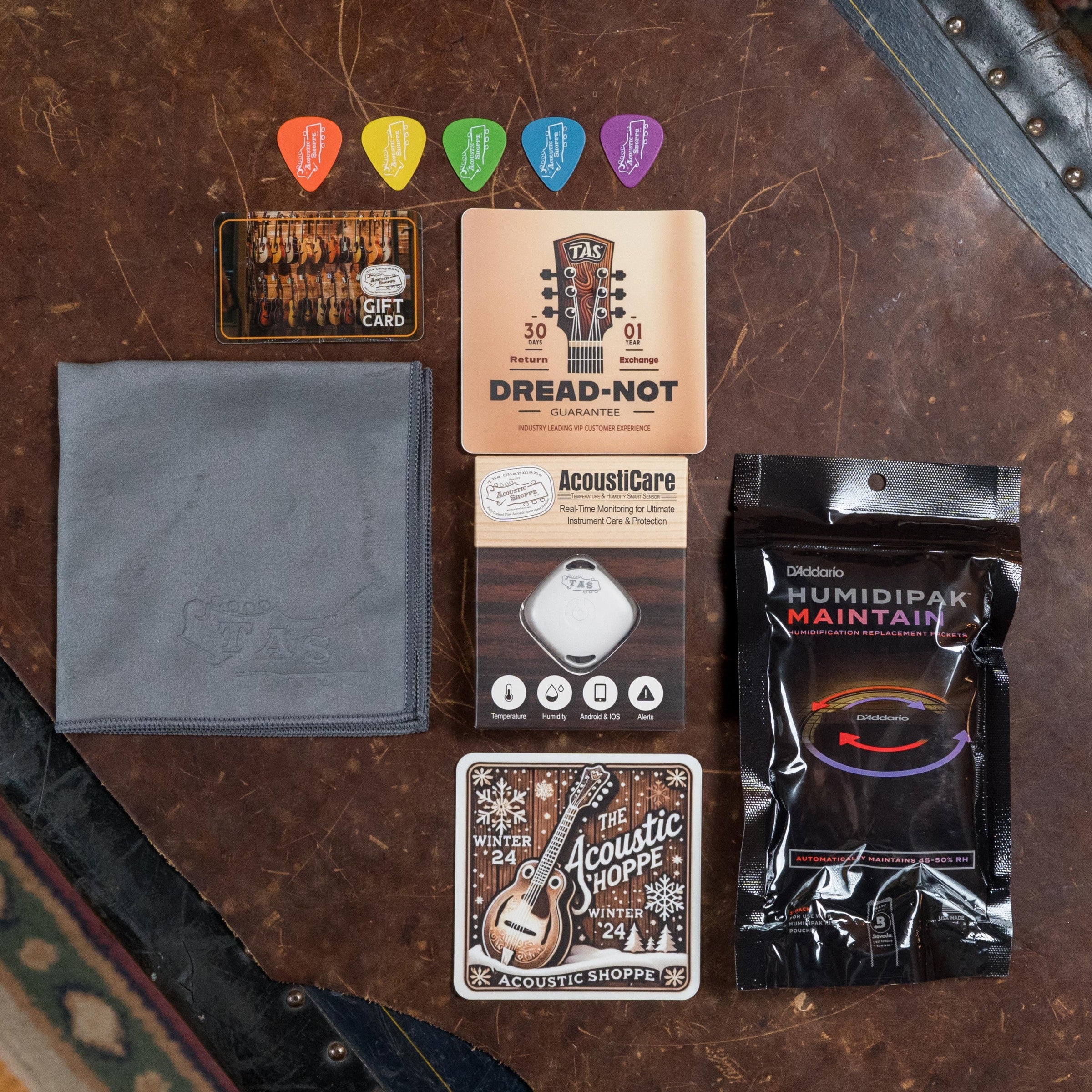 A flat lay of guitar accessories—colorful picks, a gift card, gray cleaning cloth, AcoustiCare humidifier, D’Addario Humidipak, Acoustic Shoppe coasters—plus a nod to the vintage tone of the Atkin Guitars White Rice Baked Sitka/Rosewood Dreadnought #3497.