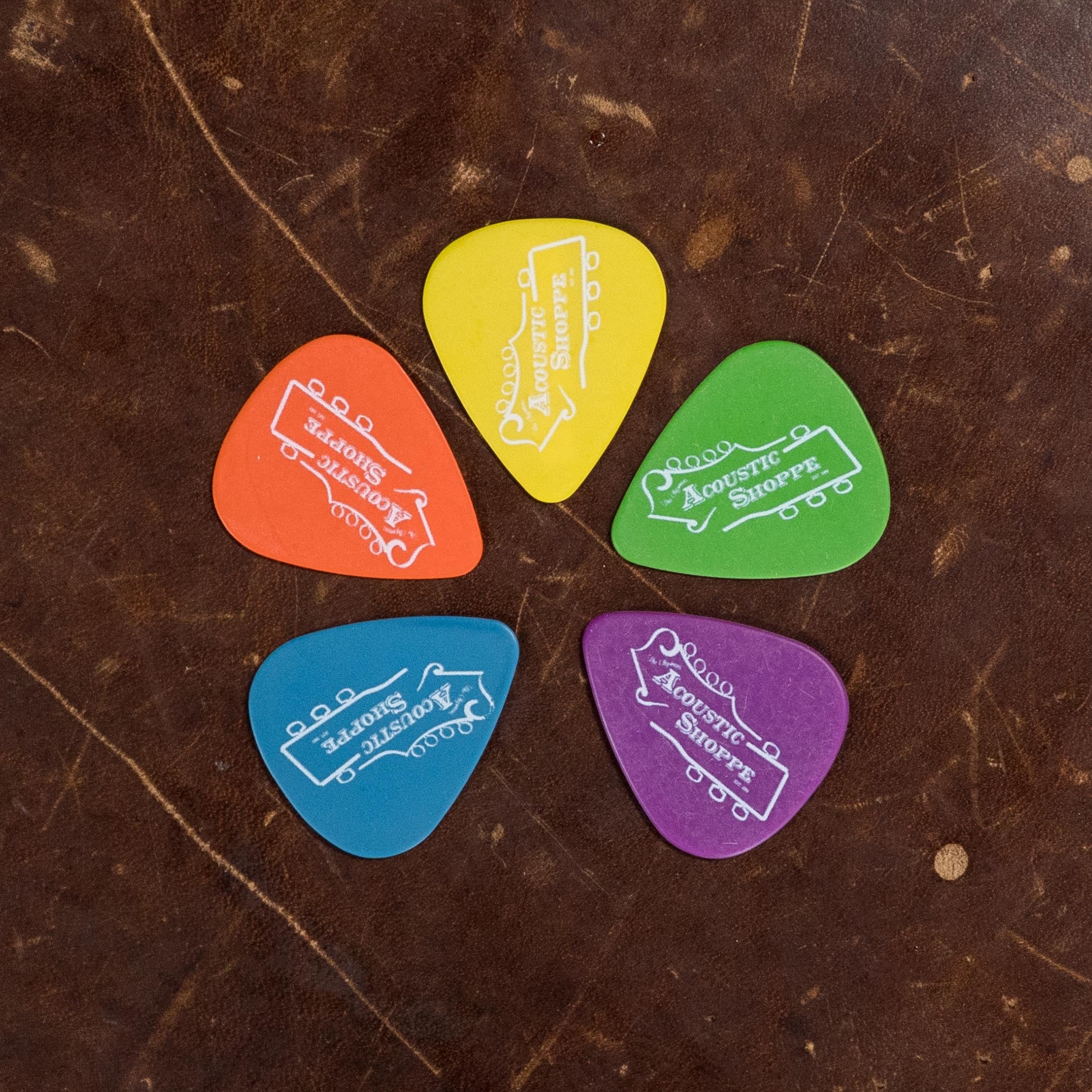 Five colorful guitar picks are arranged on worn brown leather. Each pick features the Atkin Guitars logo, matching perfectly with the Atkin White Rice Baked Sitka/Rosewood Dreadnought Acoustic Guitar #3497 for authentic vintage tone.